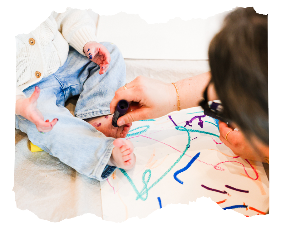 A woman drawing with colorful markers on a large sheet of paper while a young child sits nearby, touching their own leg.