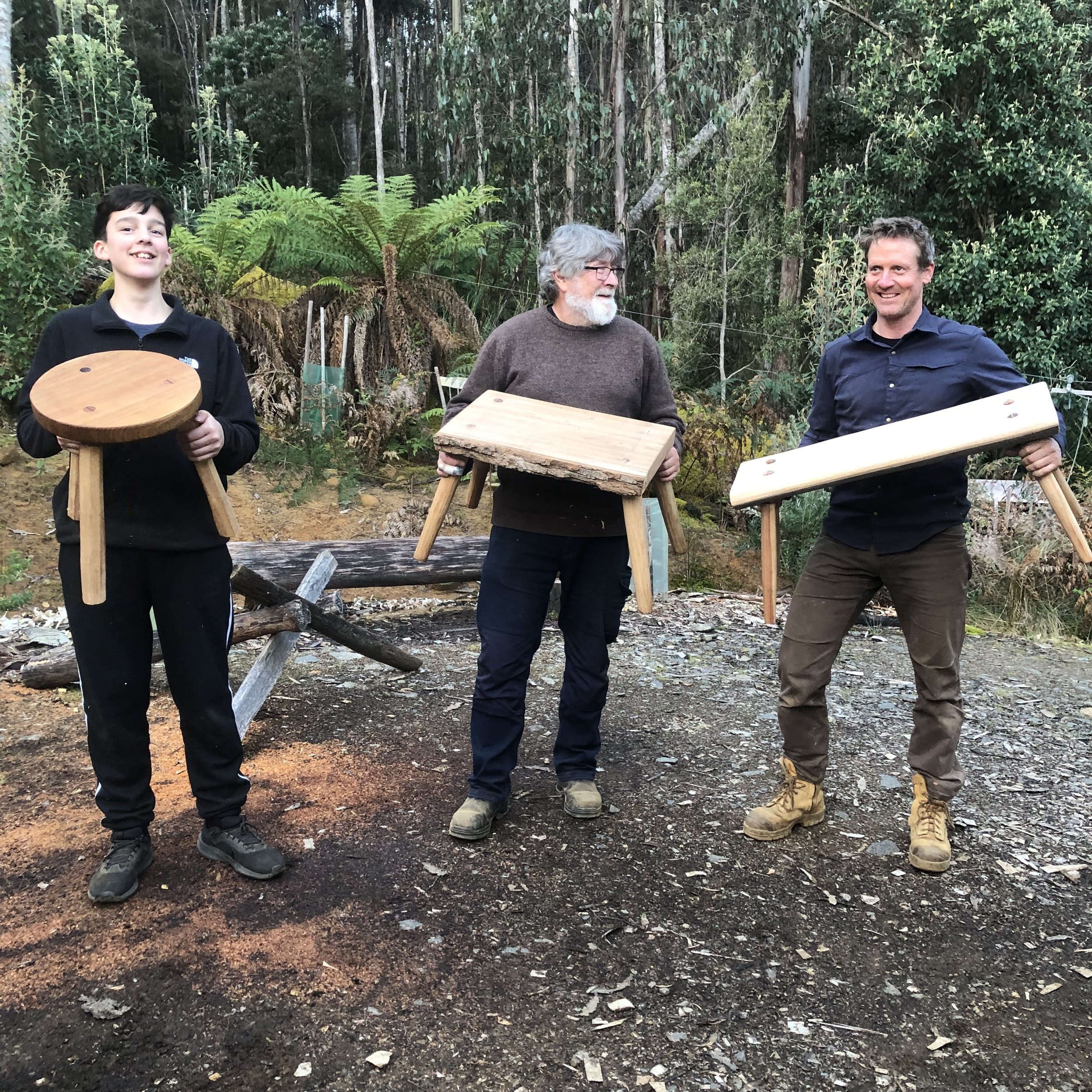 Three workshop participants holding their hand-crafted stool and benches after the 'Make Simple Furniture' course, Wisdom Through Wood.