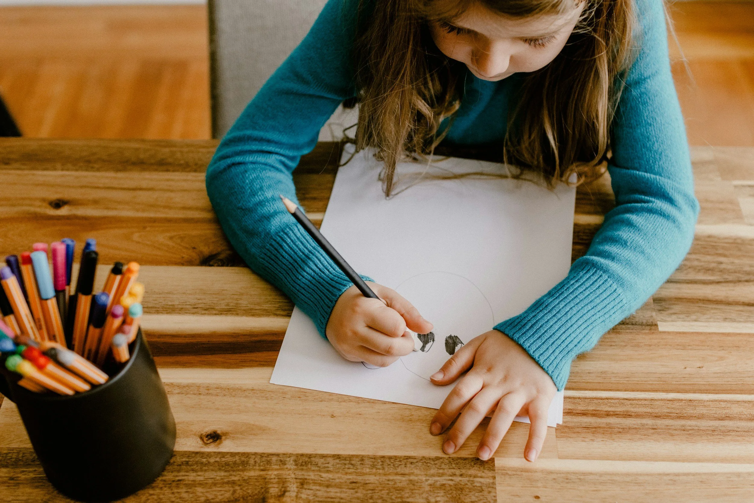 A young girl wearing a blue sweater draws on a white sheet of paper with a black pencil at a wooden table. There are colored markers in a black container on the table.