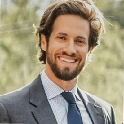 A man with dark brown hair, beard, and mustache smiling outdoors, wearing a suit jacket, dress shirt, and tie, with a blurred green nature background.