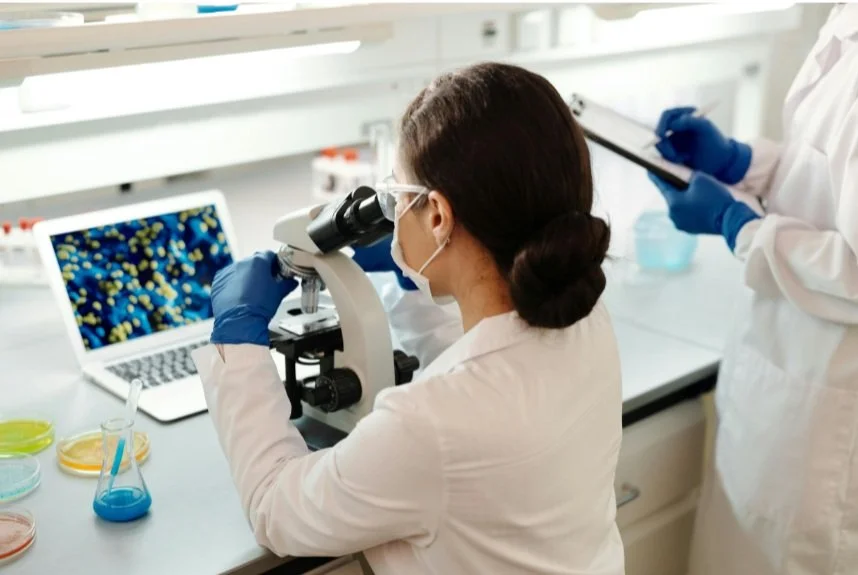 A female scientist using a microscope in a laboratory, with another scientist taking notes nearby.