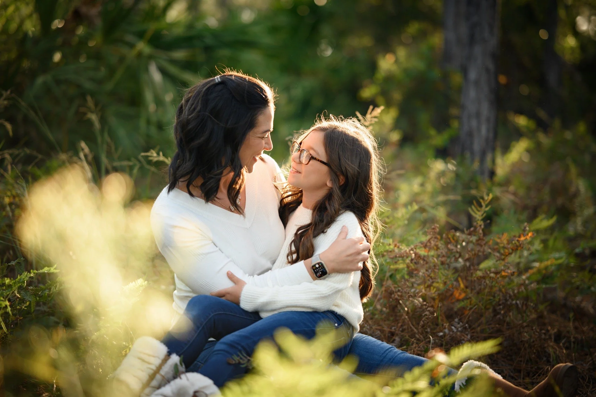 A woman and a girl sitting together outdoors among green foliage, smiling and looking at each other, embracing lovingly in warm sunlight.
