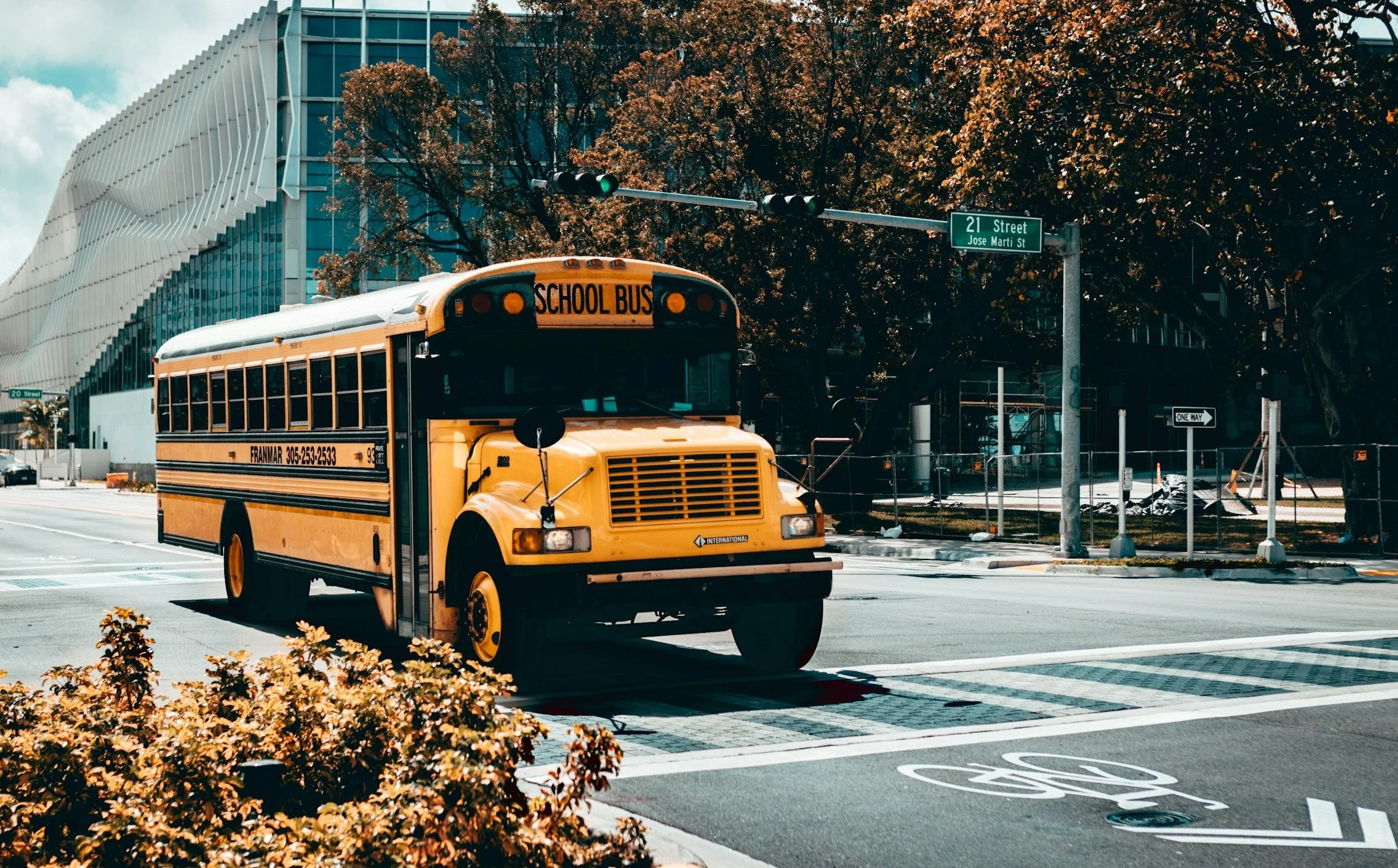 A yellow school bus crossing an urban street with modern buildings and trees with orange leaves in the background. There are street signs and a bike lane on the road.