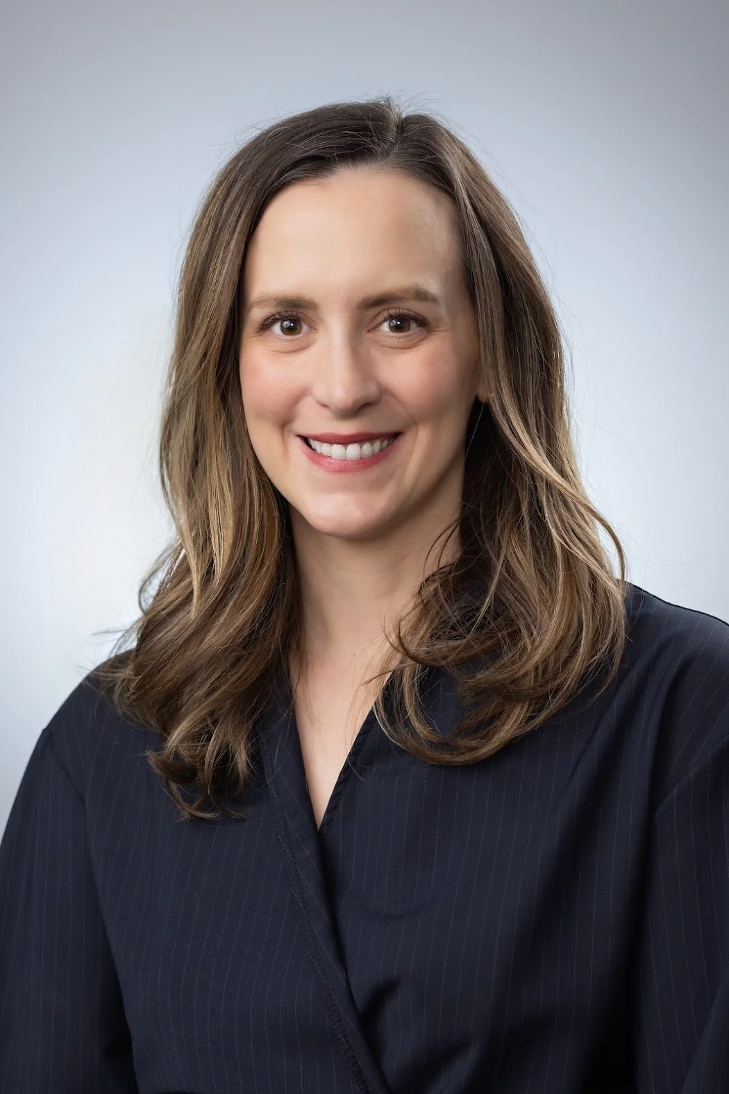 Professional woman with long, wavy brown hair smiling at the camera, wearing a dark blazer against a plain background.
