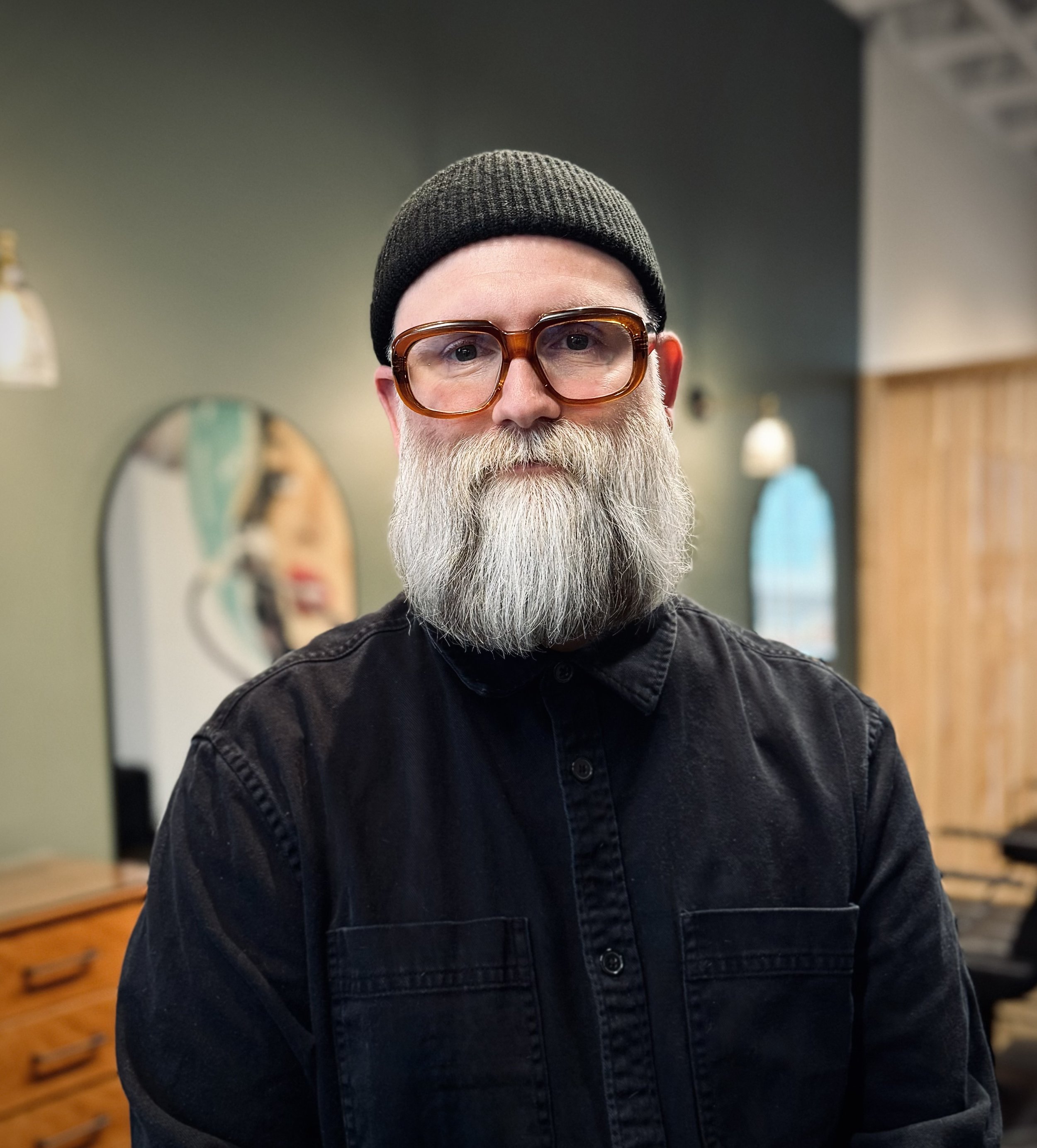 A man with a long white beard wearing large glasses, a black beanie, and a black shirt. He is standing indoors in front of a mirror and wooden furniture.