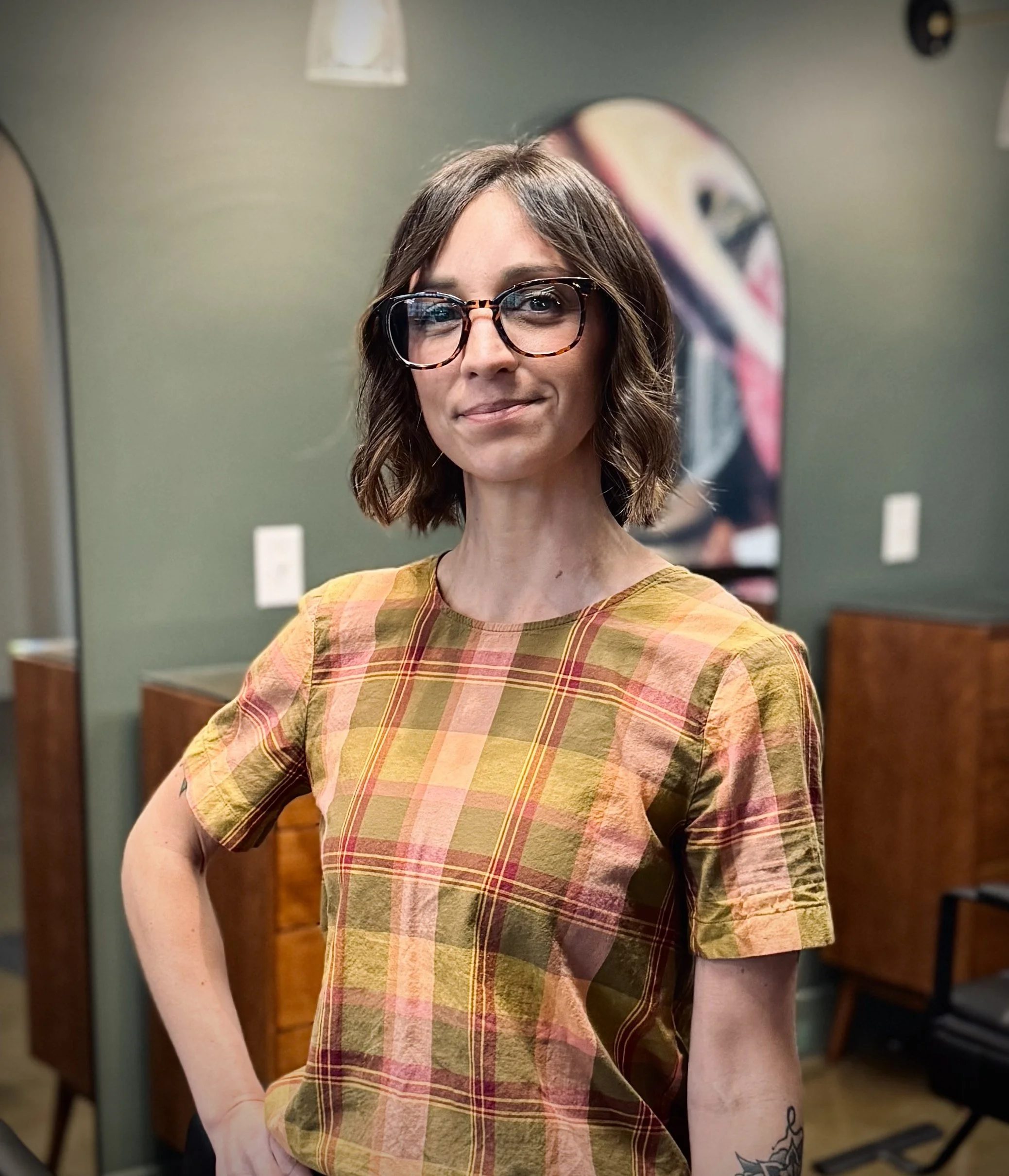 A woman with shoulder-length brown hair, glasses, and a tattoo on her left arm stands in a room with green walls and wooden furniture, wearing a plaid shirt in shades of yellow, orange, and pink.