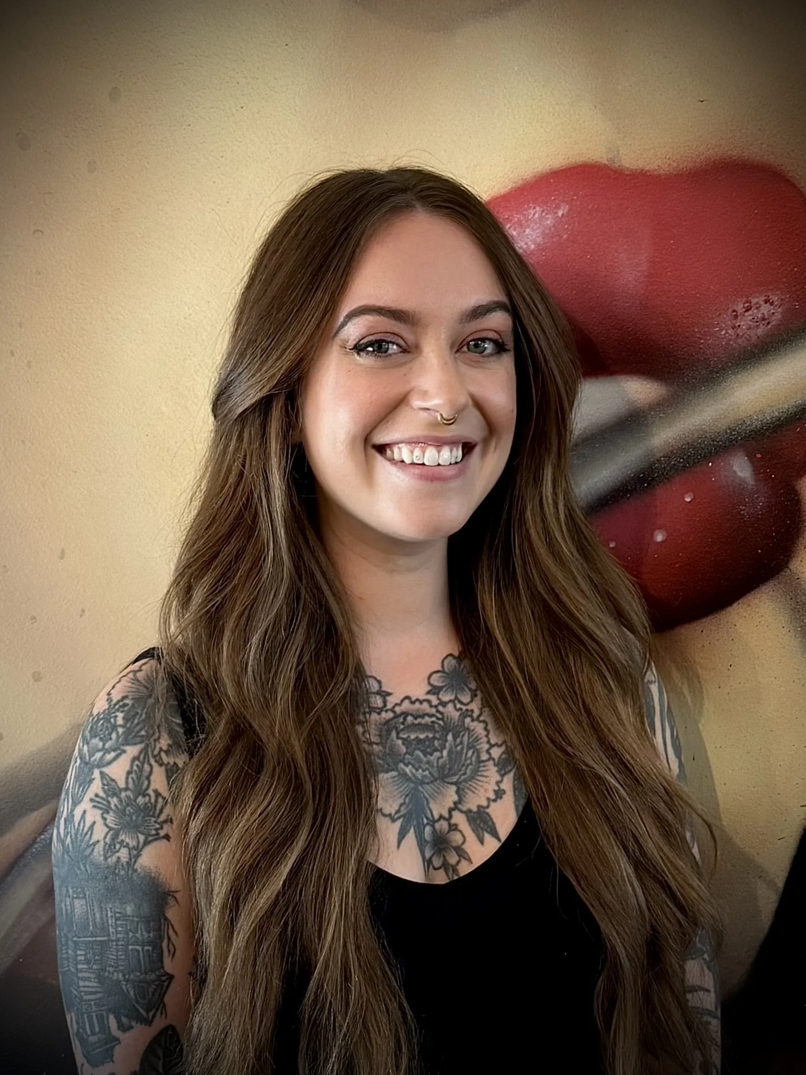 A woman with long brown hair, smiling, with tattoos on her arms and chest, wearing a black top, sitting in front of a wall with a large, close-up image of red lips with white teeth.
