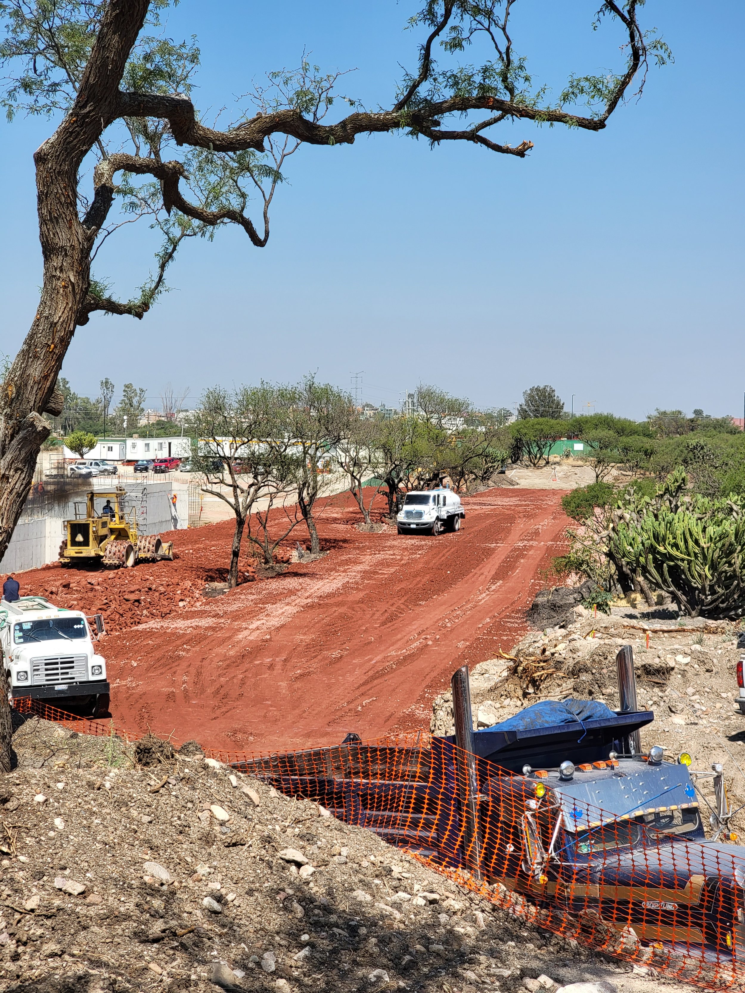 Sitio en construcción con maquinaria pesada, autos y árboles en el fondo, suelo de tierra roja y cercas de plástico anaranjado.