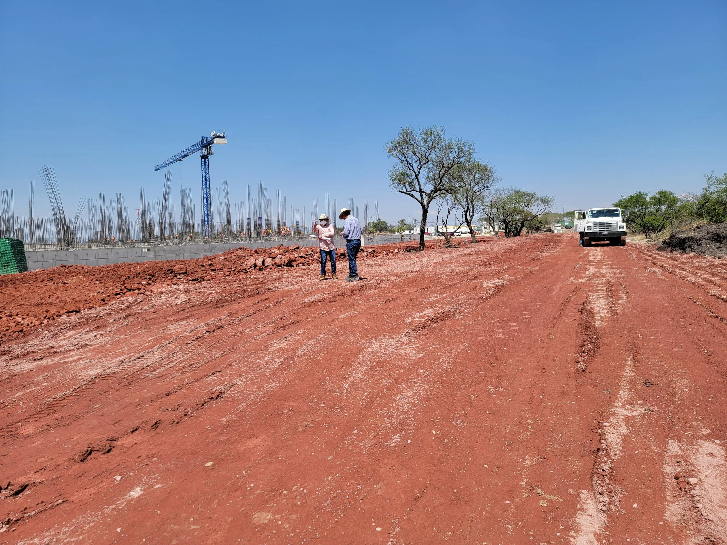 Dos personas con sombreros discuten en un camino de tierra en un lugar en construcción, con maquinaria y árboles en el fondo bajo un cielo despejado.