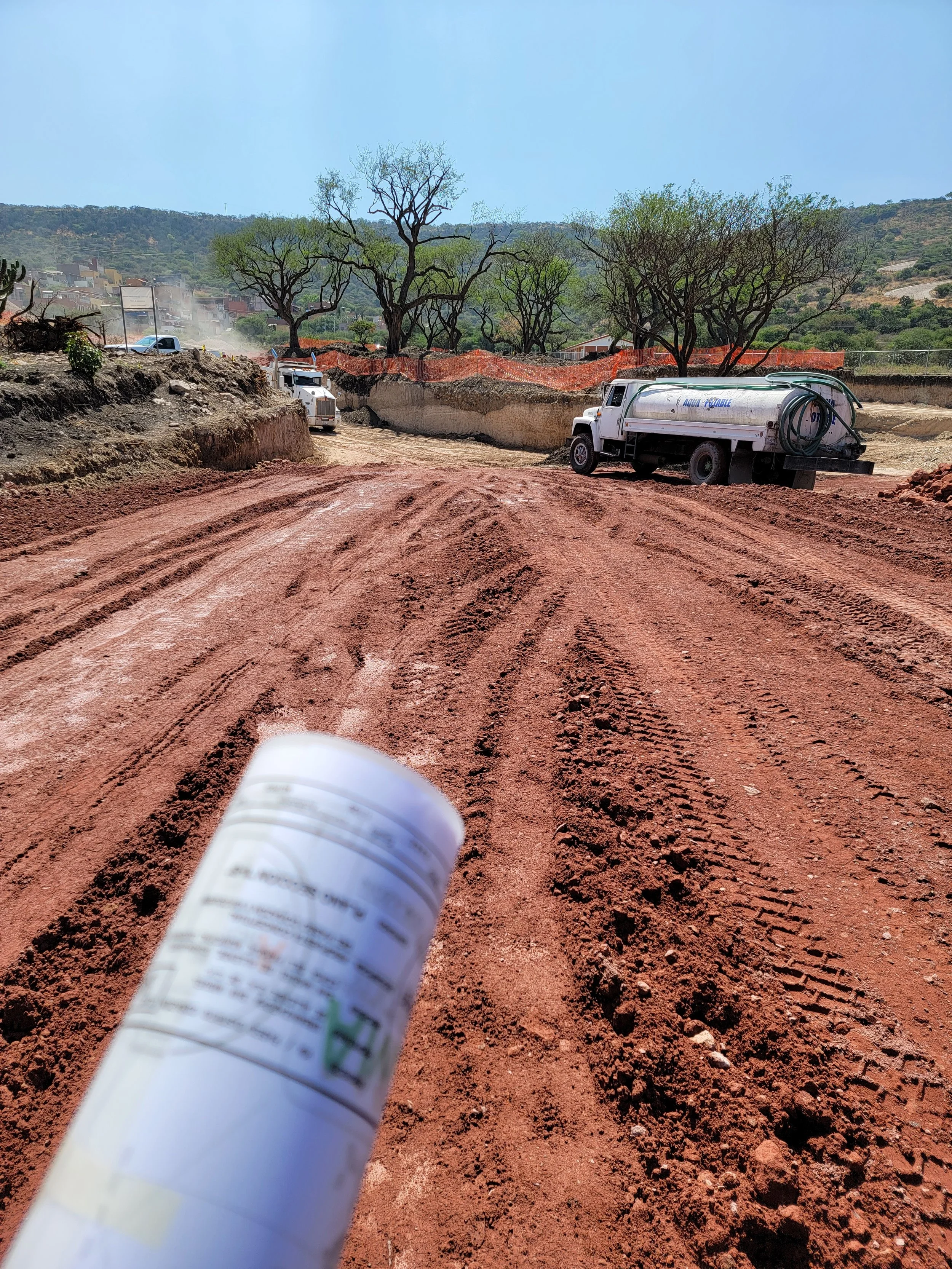 Camión cisterna en un camino de tierra en construcción, con árboles y colinas al fondo, y un envase en primer plano.