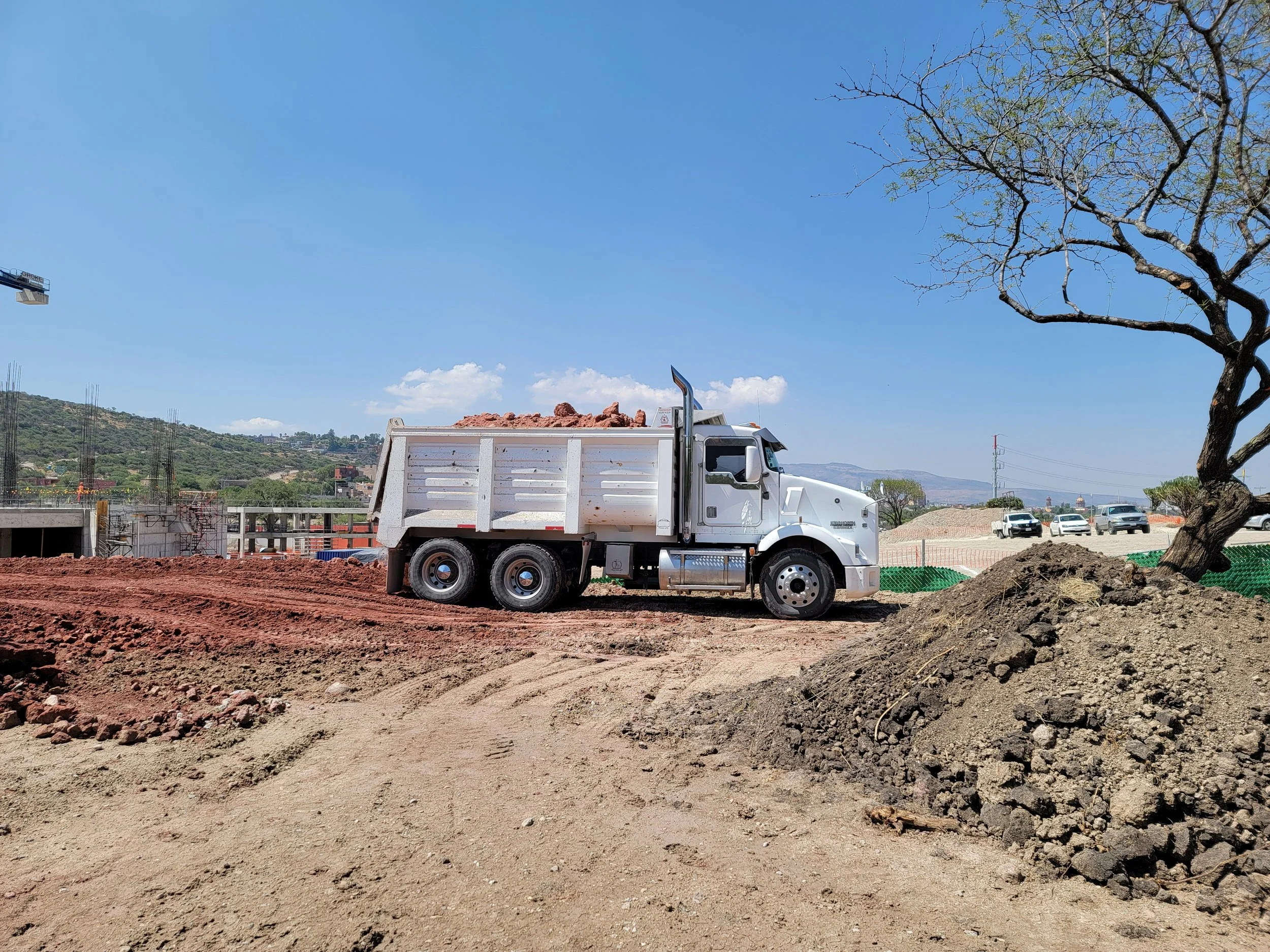 Camión de construcción en un sitio de obra con tierra y maquinaria, en un entorno con cielo despejado y algunos vehículos al fondo.