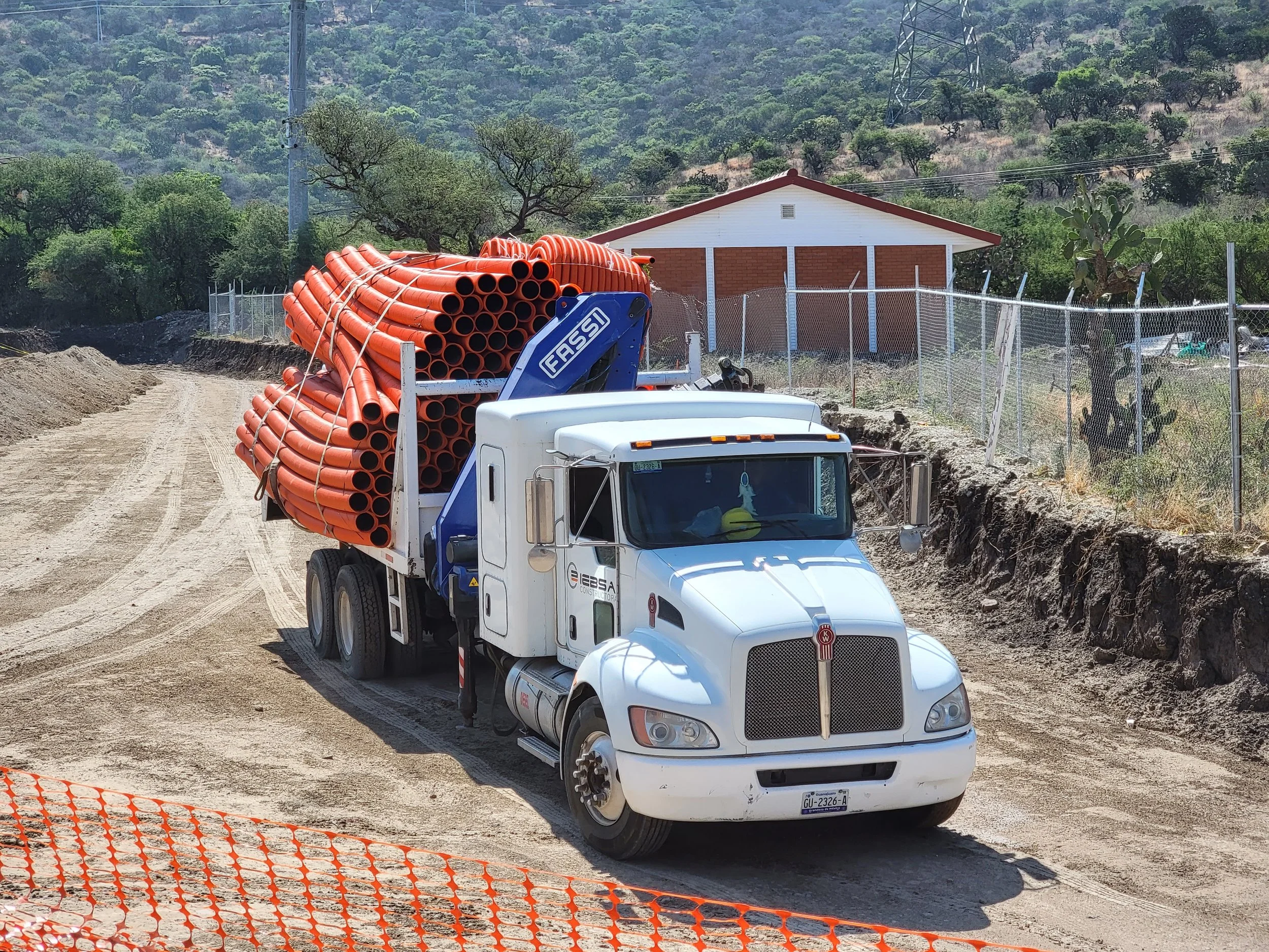 Camión de construcción con tubos de color naranja cargados en una carretera de tierra, en un entorno rural con vegetación y una casa de fondo.