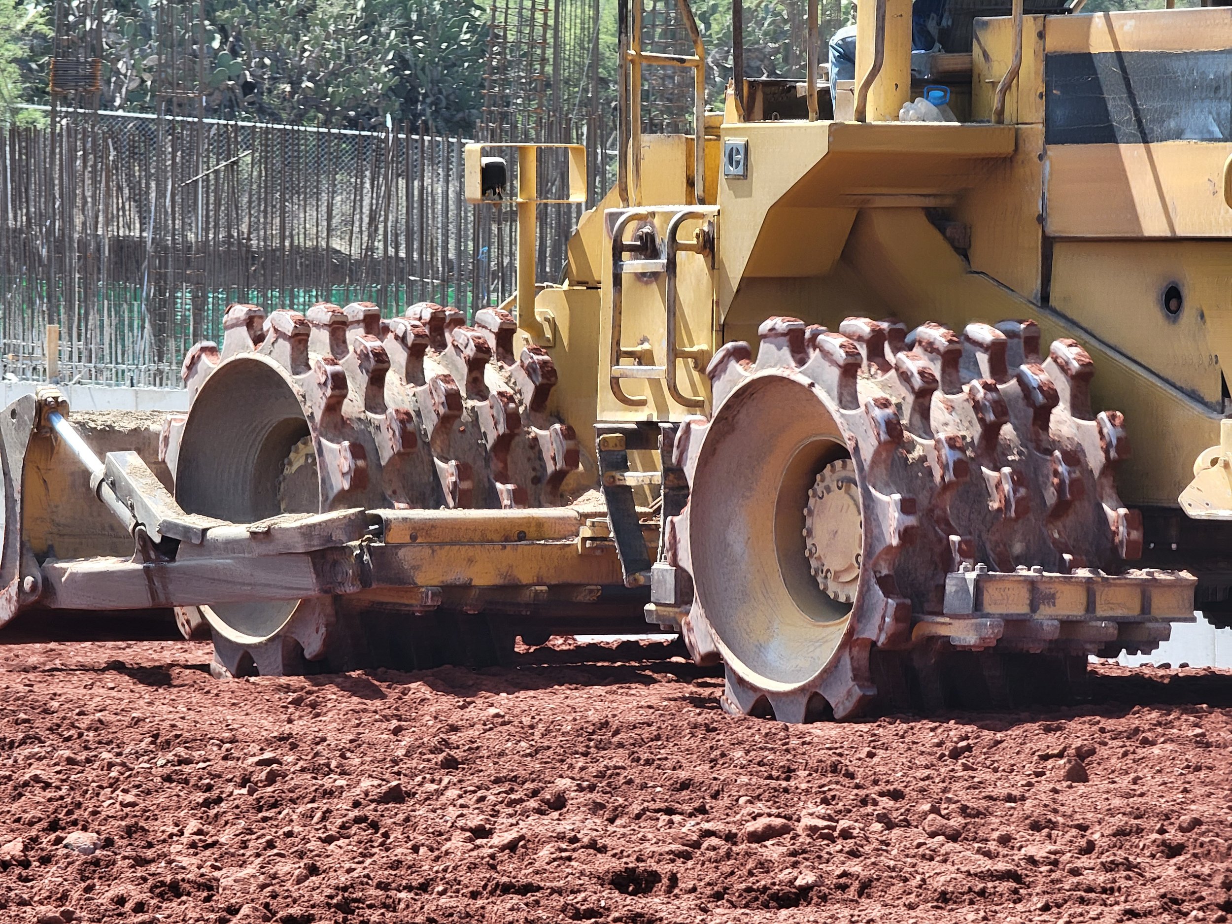 Maquinaria de construcción amarilla en proceso de compactación de tierra con superficie de tierra roja y fondo con árboles y una cerca de madera.