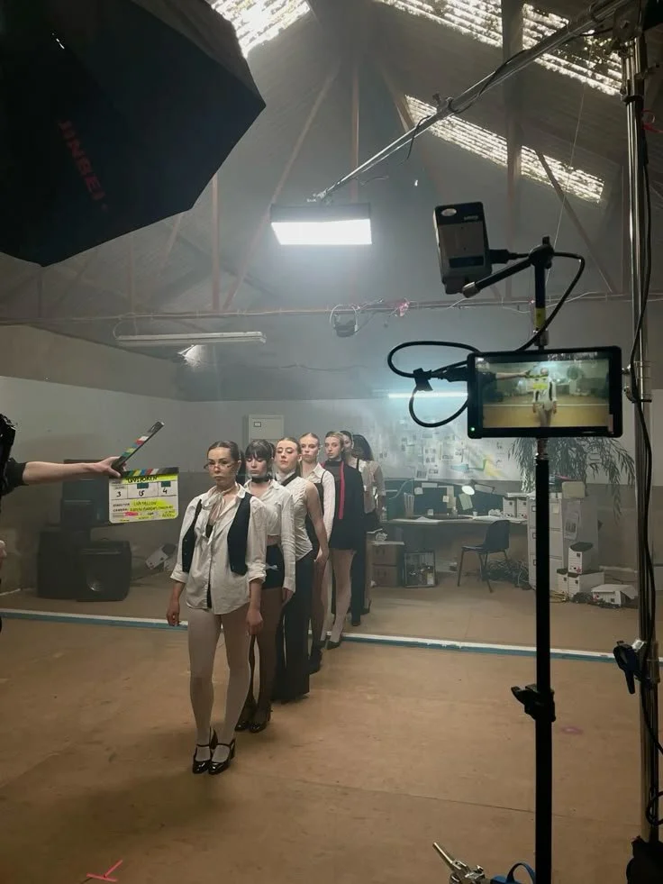 Grupo de modelos femininas posando para uma gravação ou sessão fotográfica em um estúdio, com equipamentos de iluminação e câmeras ao redor.