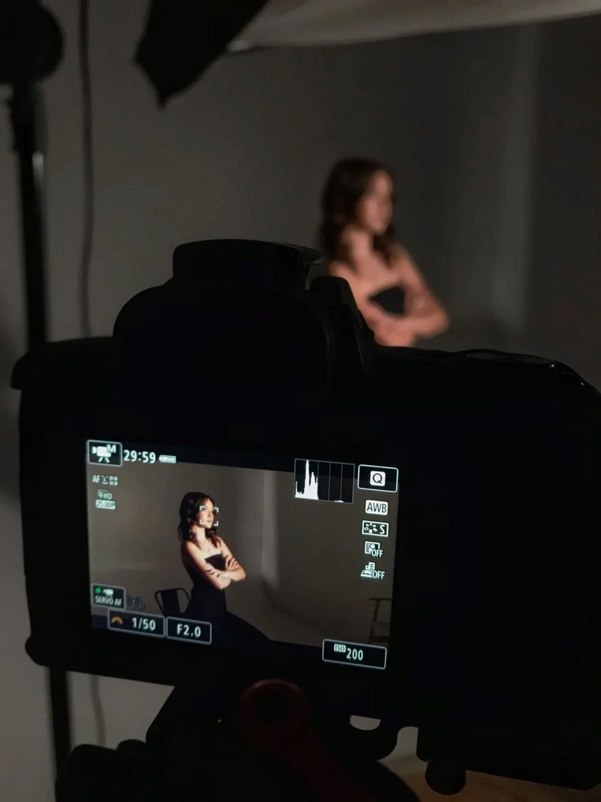 Foto de uma câmera fotográfica voltada para uma mulher com braços cruzados, usando vestido preto, em um estúdio com fundo cinza. O visor da câmera mostra a mesma mulher, com uma postura similar, em uma pose de ensaio fotográfico.