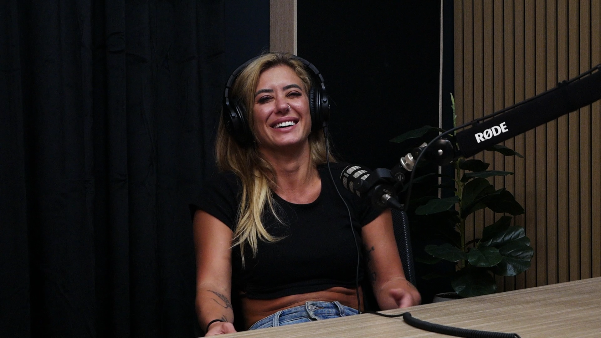 A woman with blonde hair smiling, wearing headphones, sitting in a recording studio with a microphone and a plant in the background.