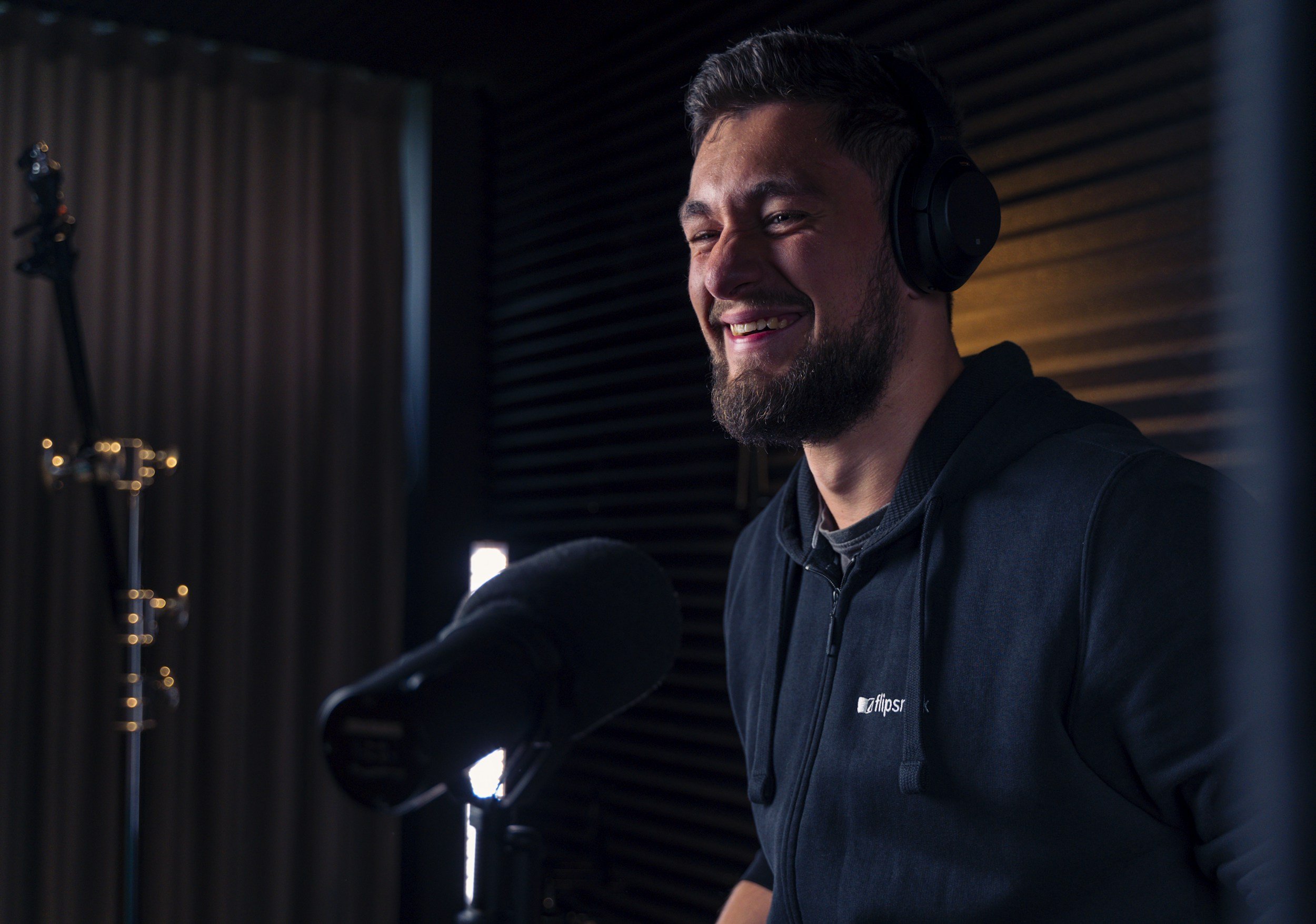 A man with dark hair and a beard wearing headphones, smiling while speaking into a microphone in a recording studio.