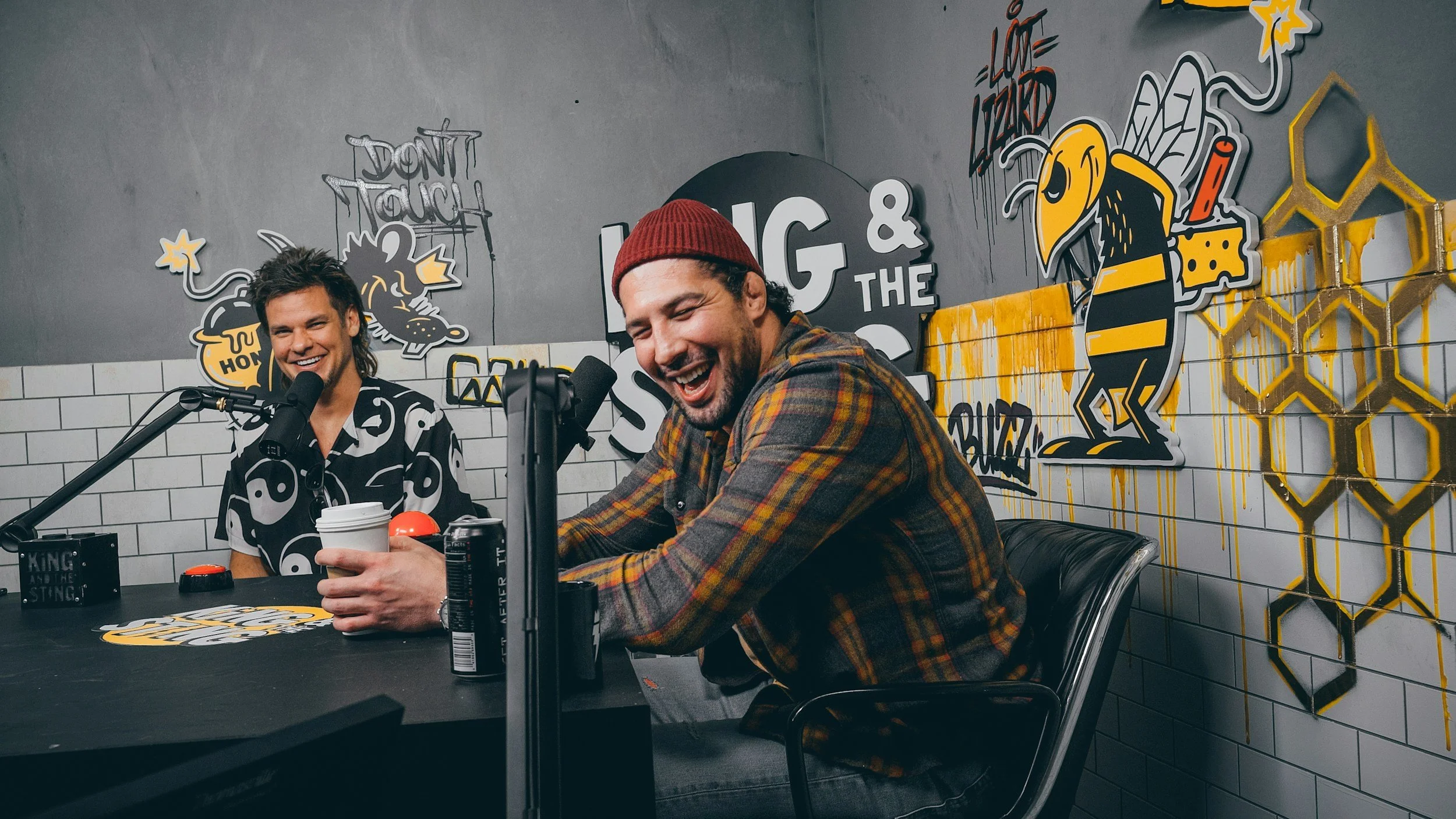 Two men laughing in a podcast studio with graffiti-style art and signs on the walls.