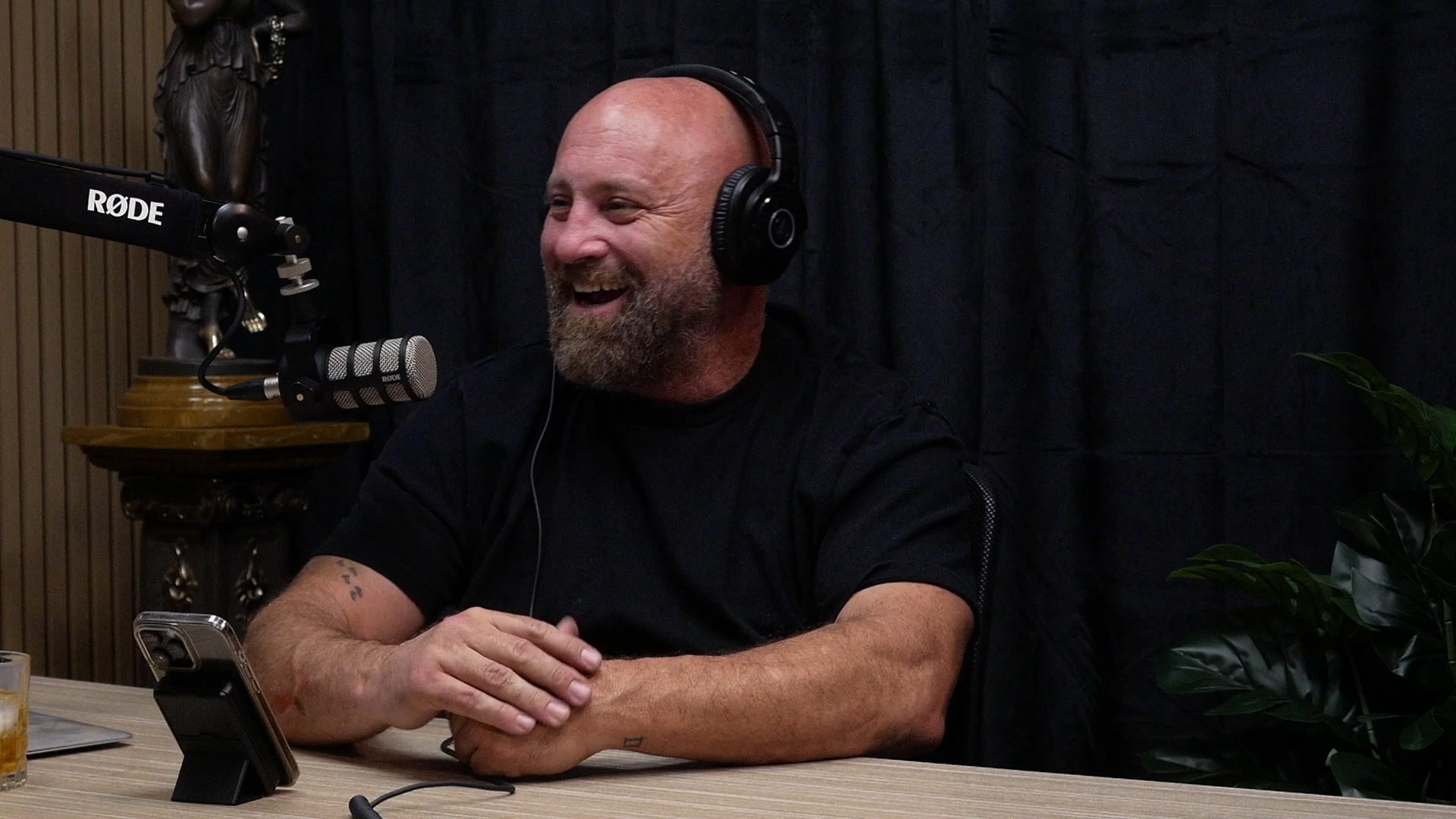 A man with a bald head and beard, wearing black headphones, smiling and sitting at a table with a microphone, a phone, and a glass of drink.