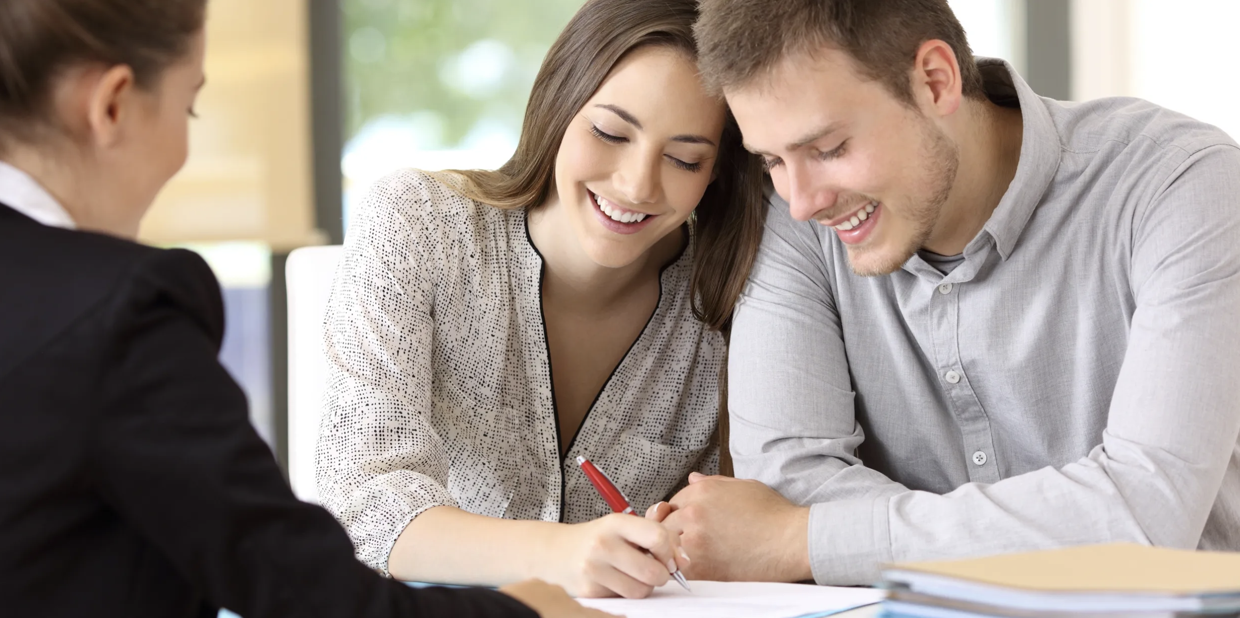 A smiling woman and man sitting at a table, signing documents, with a person on the left side of the image.