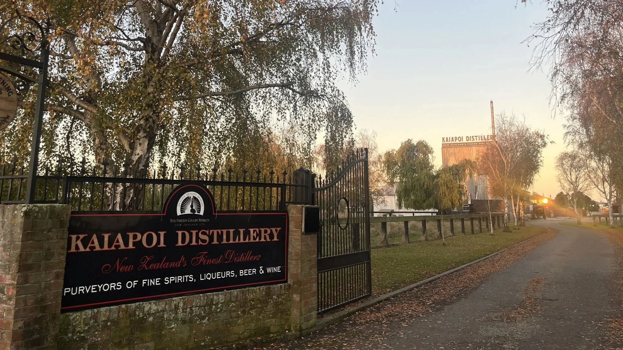 Entrance to Kaiapoi Distillery with a sign reading 'Purveyors of fine spirits, liqueurs, beer & wine' and a building in the background, surrounded by trees during sunset.