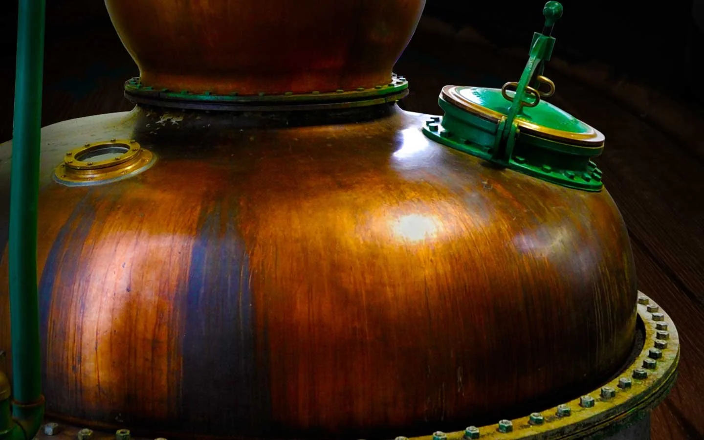 Close-up of an industrial copper boiler with a green lid and green pipe, set on a dark wooden floor.