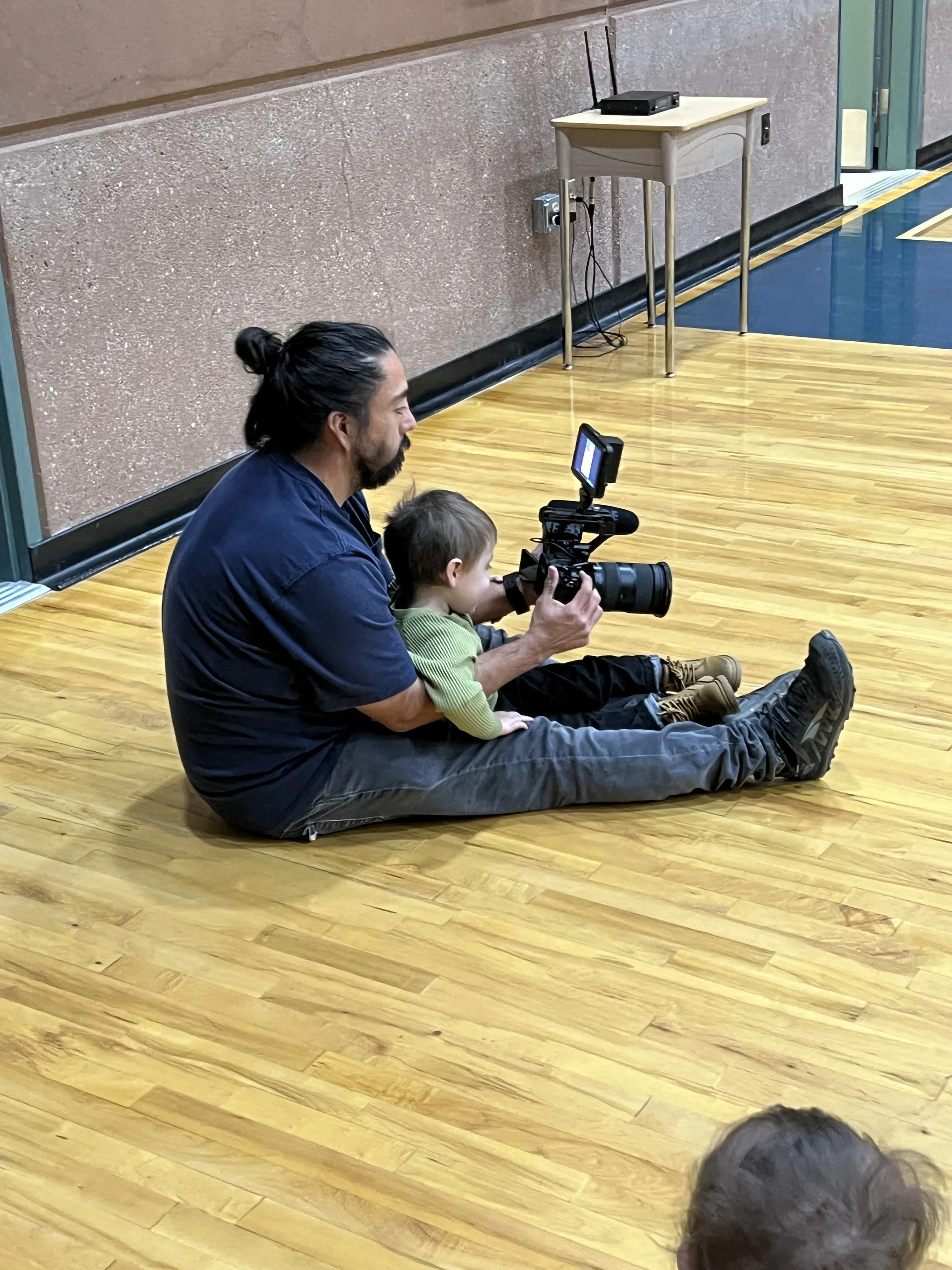 A man with long black hair tied in a bun and a beard sitting on a wooden floor, holding a camera with a young boy sitting on his lap, both looking through the camera viewfinder. The background shows a pinkish wall, a small table with a router on top, and some electrical cords.