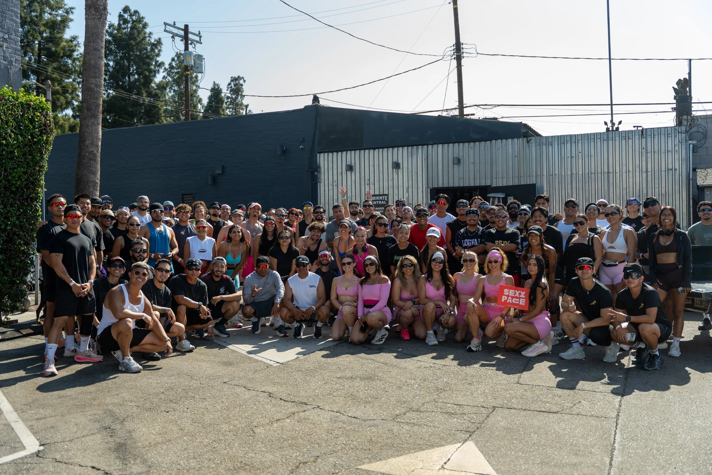 A large group of people gathered outdoors in front of a building, many wearing athletic clothing and sunglasses, with some holding a red sign that reads "SEXY PACE".