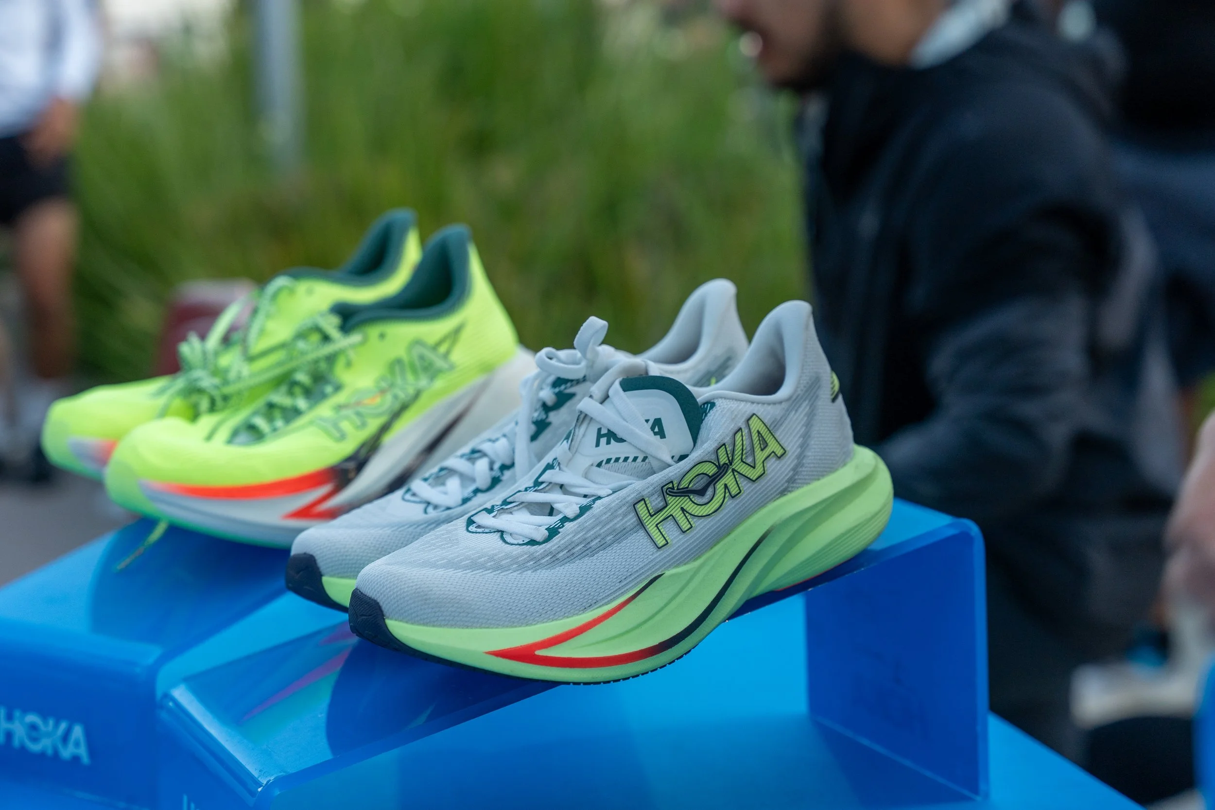 Close-up of three pairs of running shoes displayed on a blue table outdoors, with a person in dark clothing in the background.