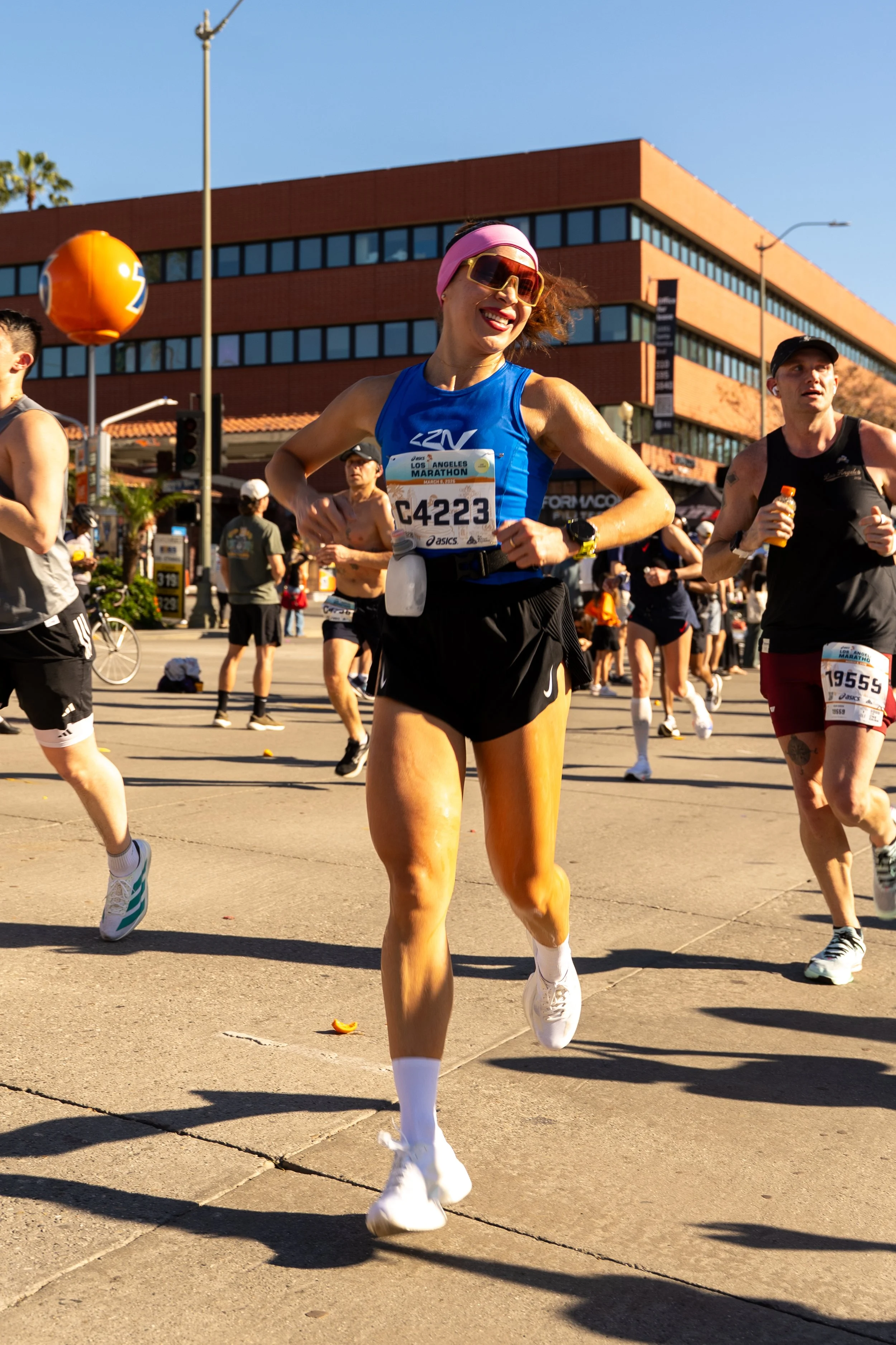 A woman running in a marathon, wearing a blue athletic top, black shorts, white running shoes, pink sunglasses, pink headband, and a yellow watch, with a bright smile on her face.