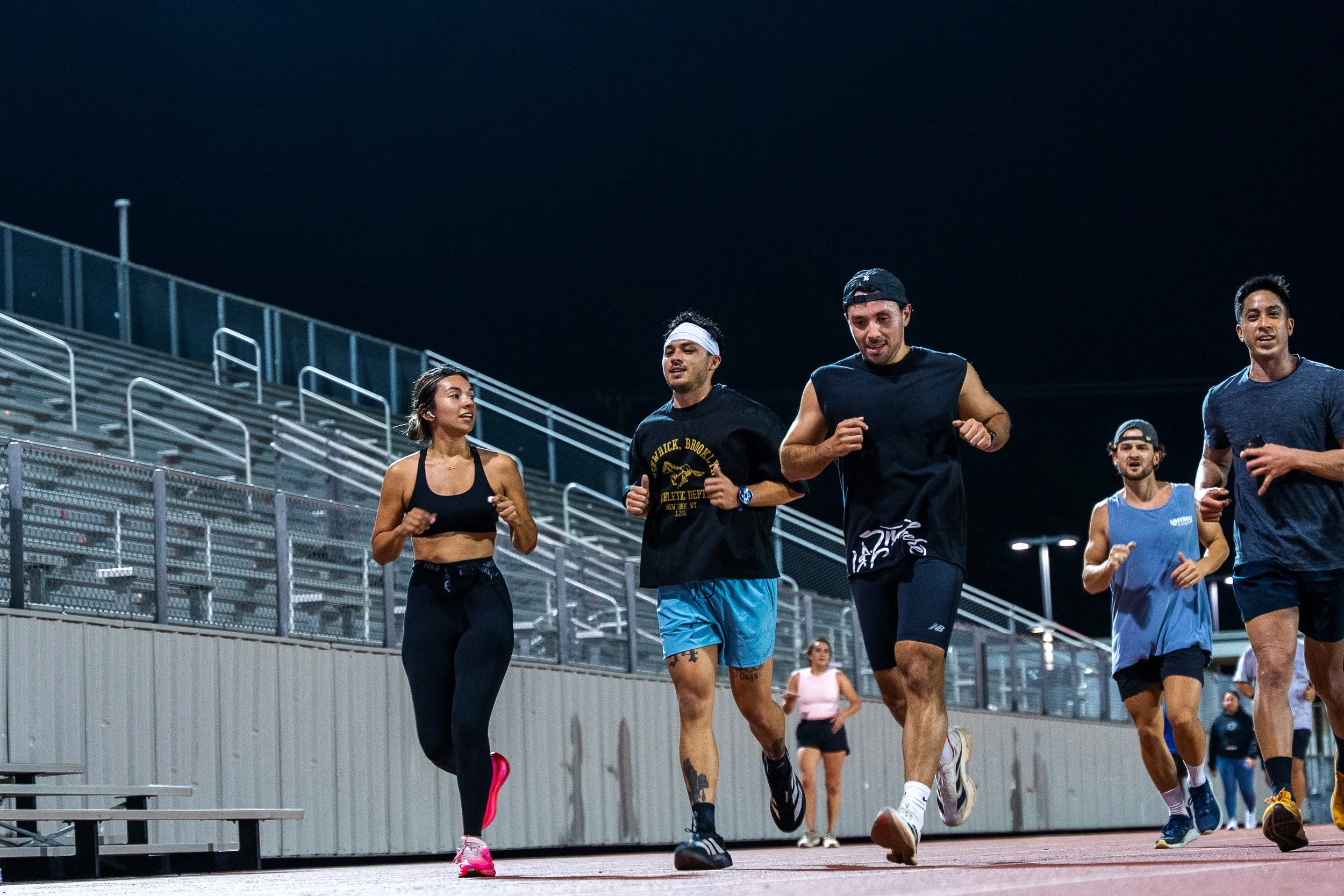 Group of people running on a track at night, with bleachers and a fence in the background.