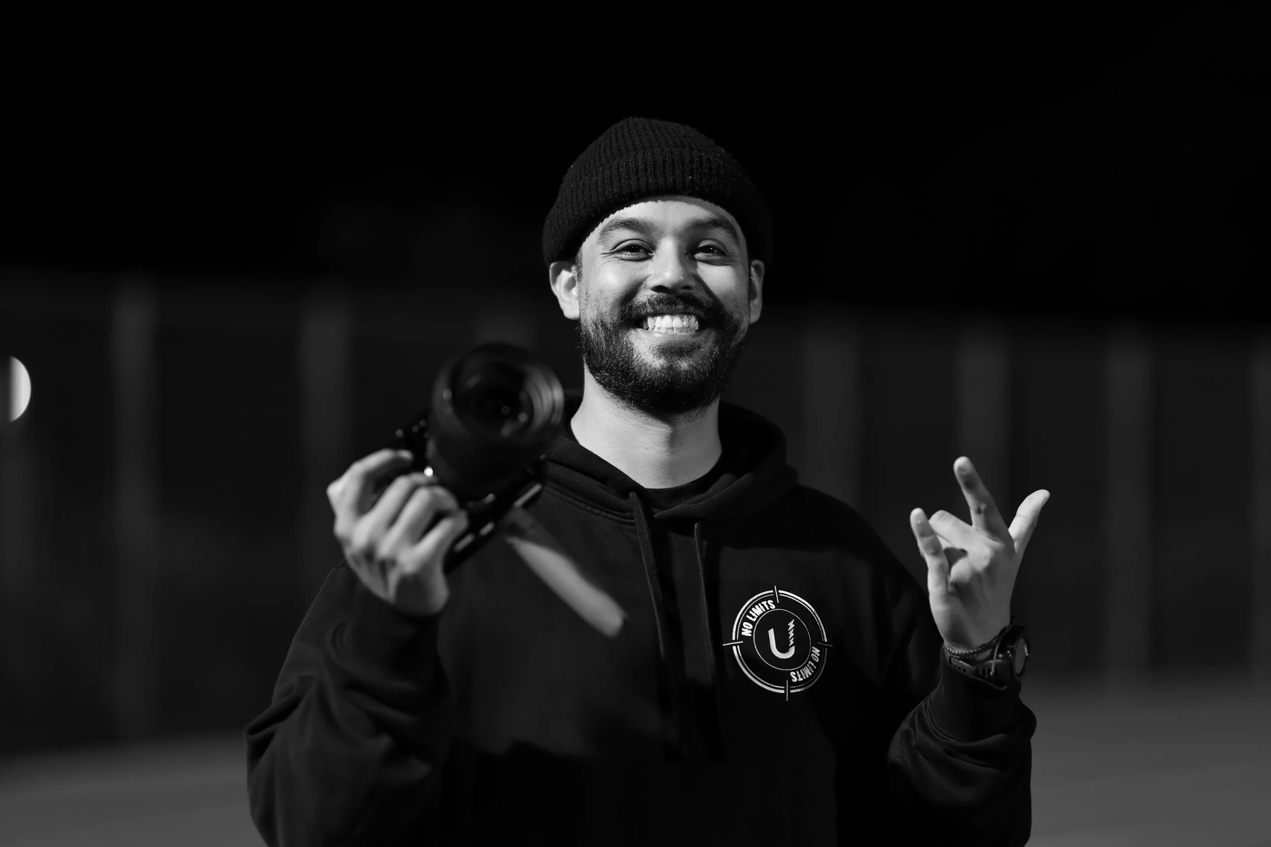 A smiling man wearing a beanie and hoodie, holding a camera, making a peace sign with his hand, against a dark background.