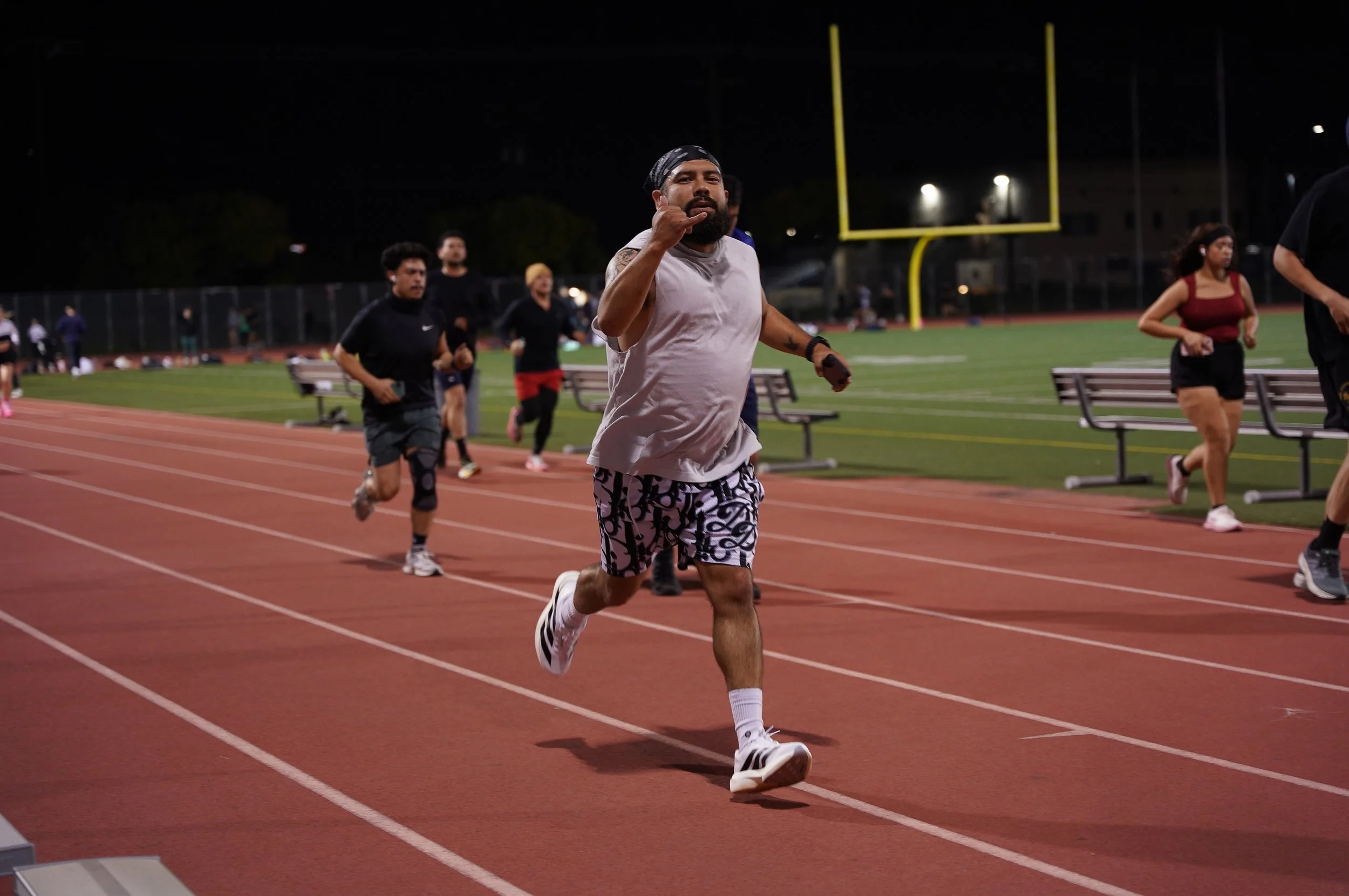 A man running on a track at night during a race, making a hand gesture and surrounded by other runners with a football goalpost in the background.