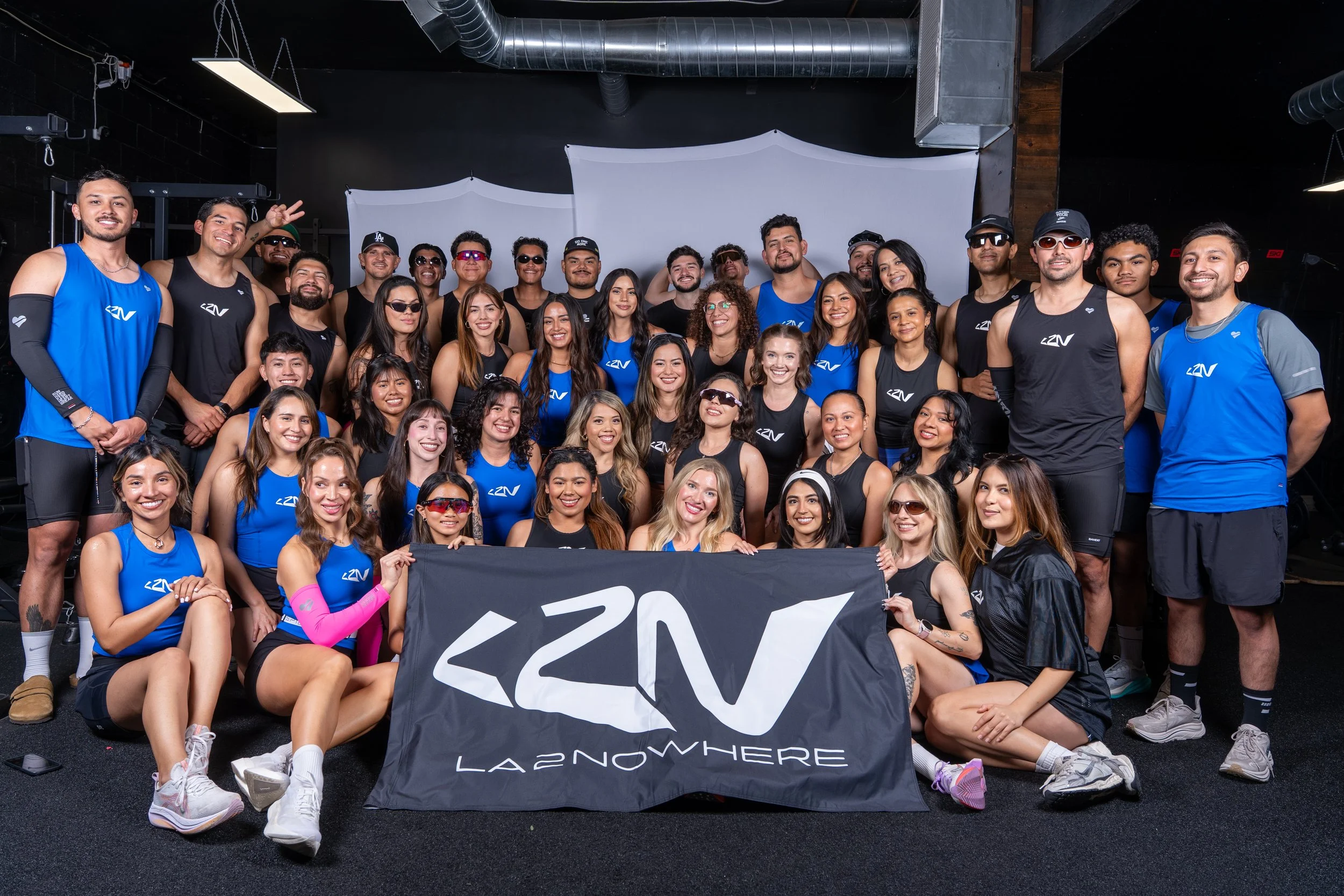 Group of diverse people in athletic clothing, smiling and posing indoors in front of a white backdrop, holding a banner that reads 'LA2NOWHERE', with some wearing sunglasses and others making peace signs.