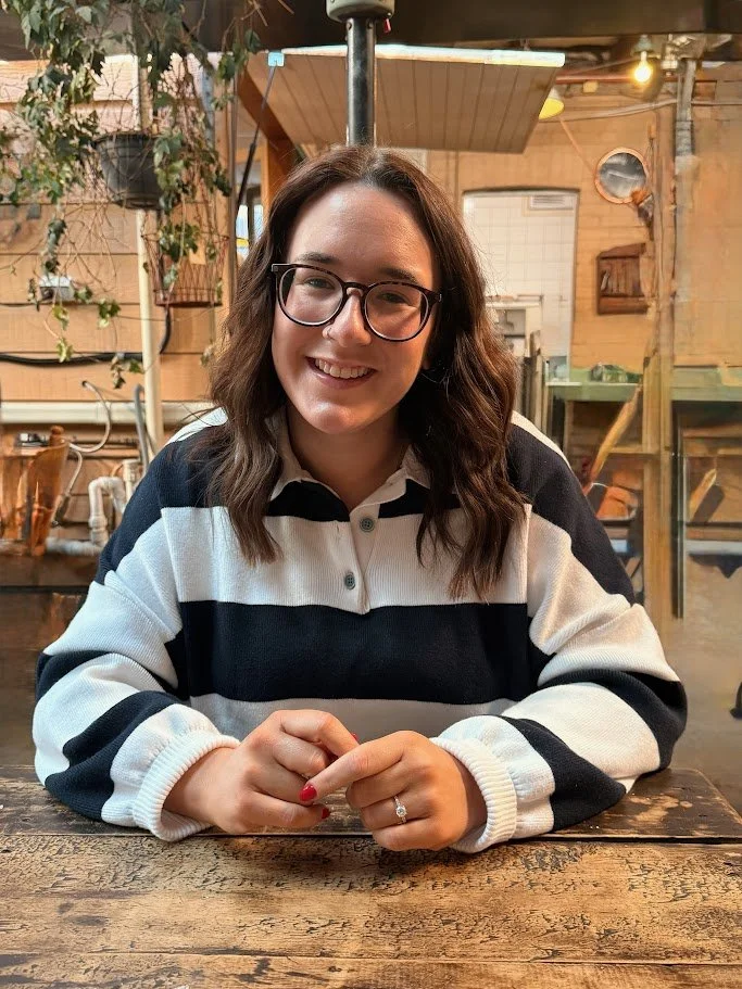 A smiling woman with glasses and wavy brown hair sitting at a wooden table in a rustic cafe or restaurant with wooden walls, plants, and warm lighting.