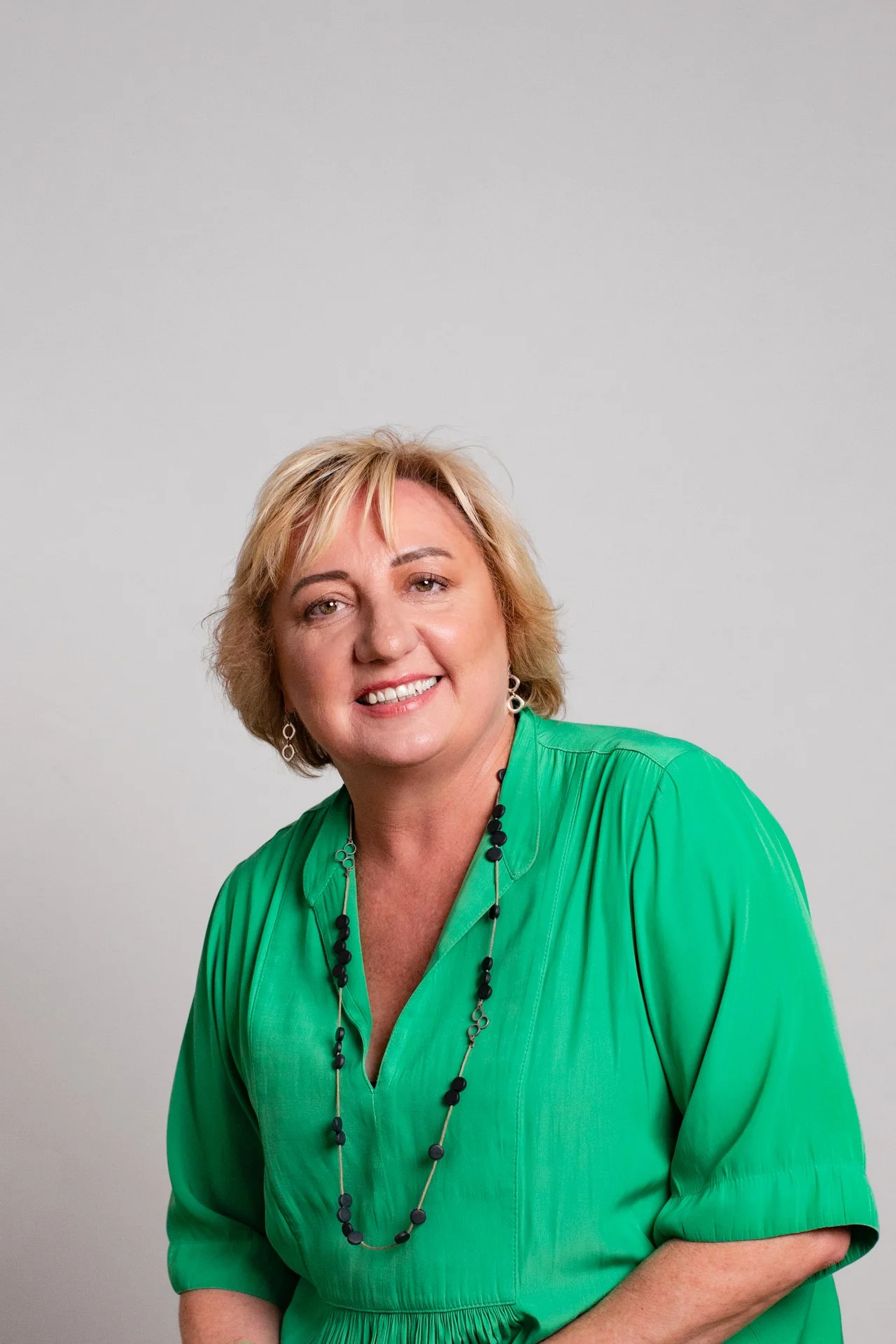 Portrait of a smiling woman with blonde hair, wearing a green dress and a black beaded necklace, against a plain light gray background.