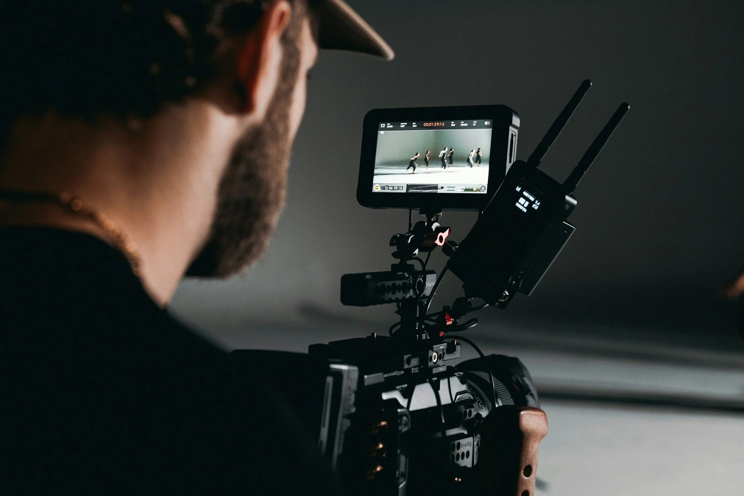 A man wearing a hat operates a professional video camera on a tripod to film a dance performance, with a monitor displaying the dancers.