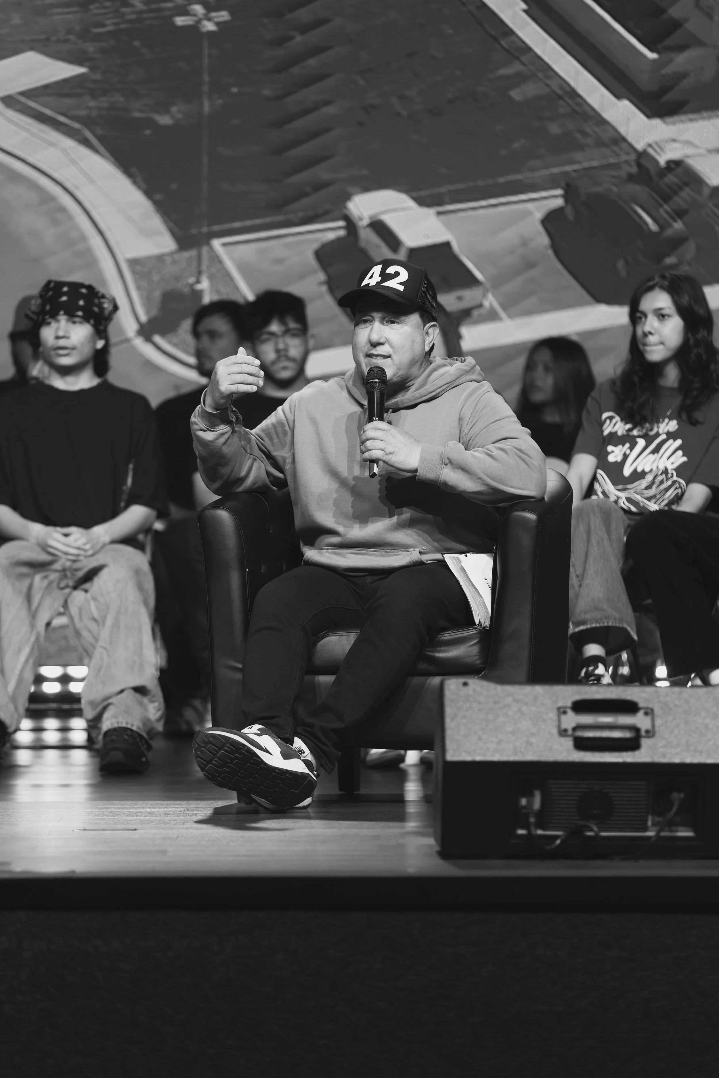 A man wearing a baseball cap with the number 42, hoodie, and sneakers speaking into a microphone while seated on stage, with people sitting behind him.