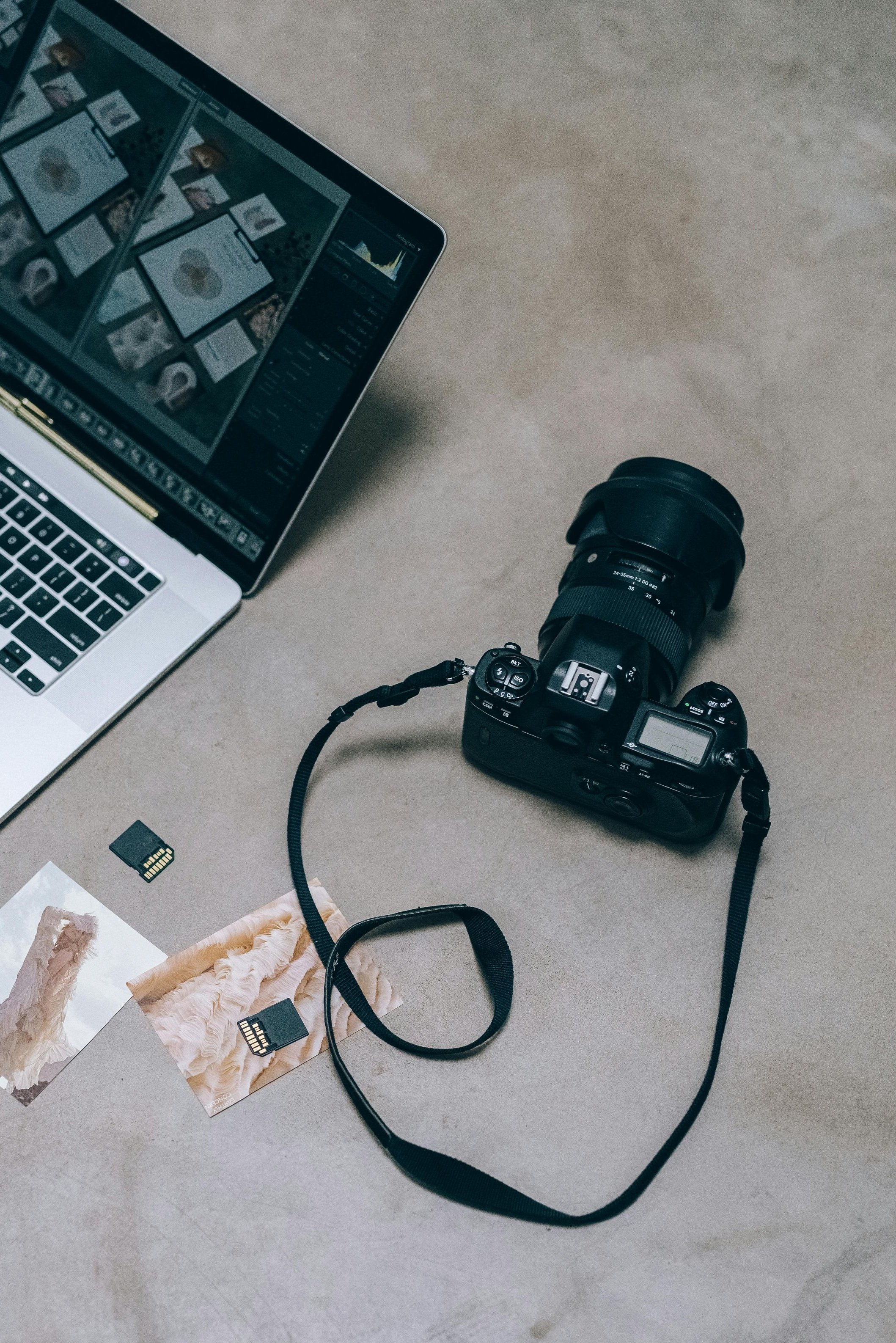 A digital camera connected to a laptop computer on a beige surface, with SD cards and photographs nearby.