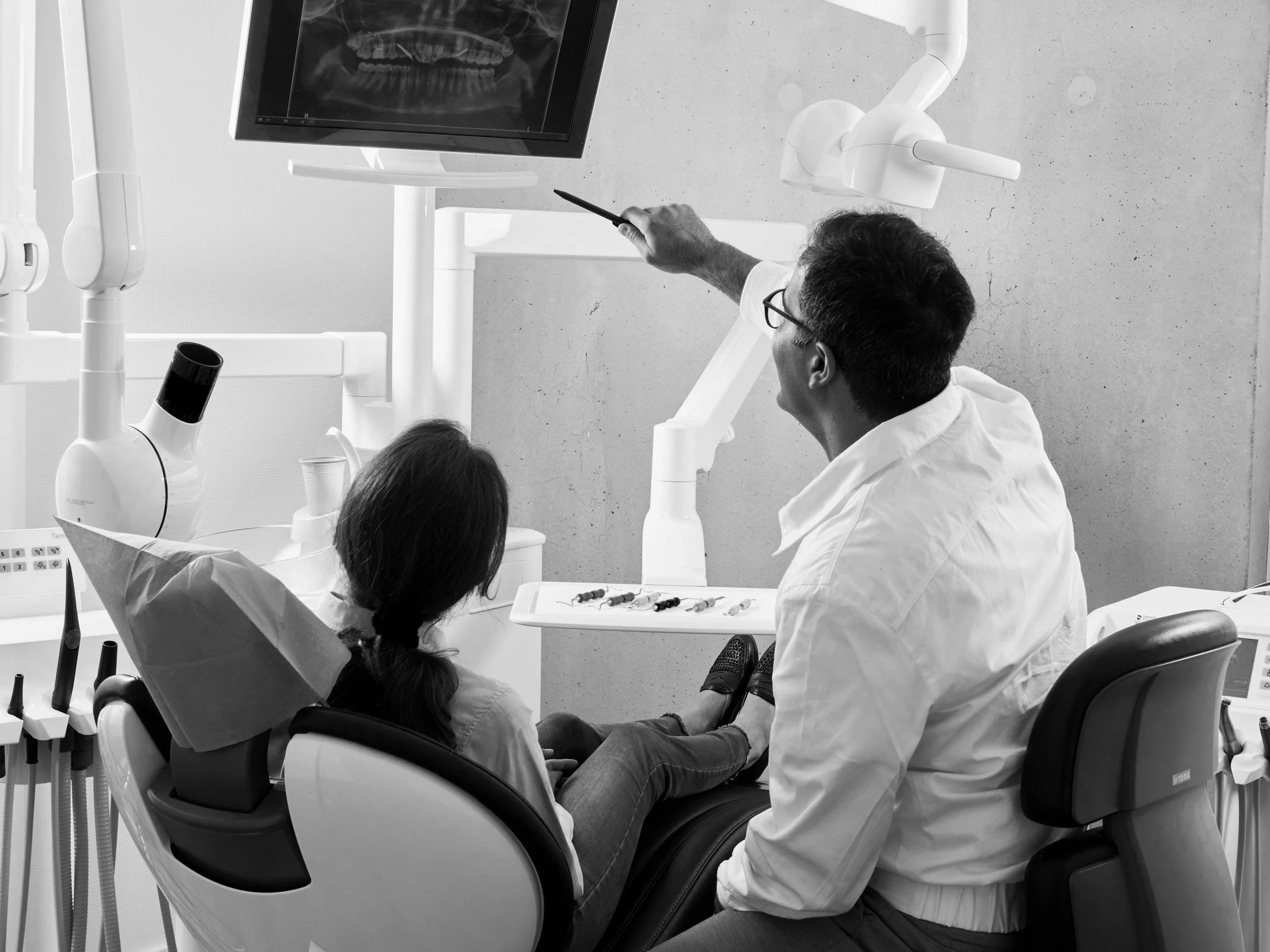 A dentist shows an X-ray on a computer screen to a young girl sitting in a dental chair. The girl is wearing a protective bib and has her feet on the lap of the dentist.