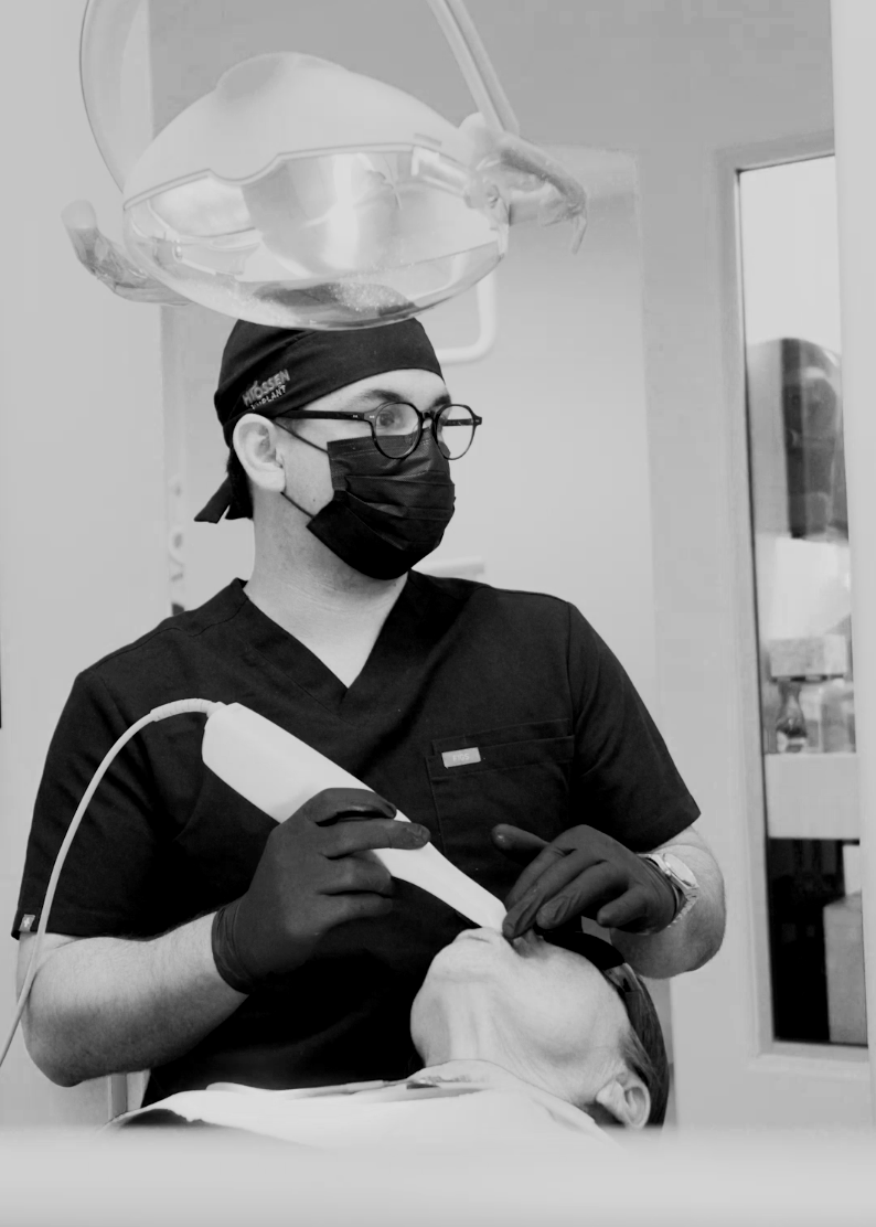 A dentist shows an X-ray on a computer screen to a young girl sitting in a dental chair. The girl is wearing a protective bib and has her feet on the lap of the dentist.
