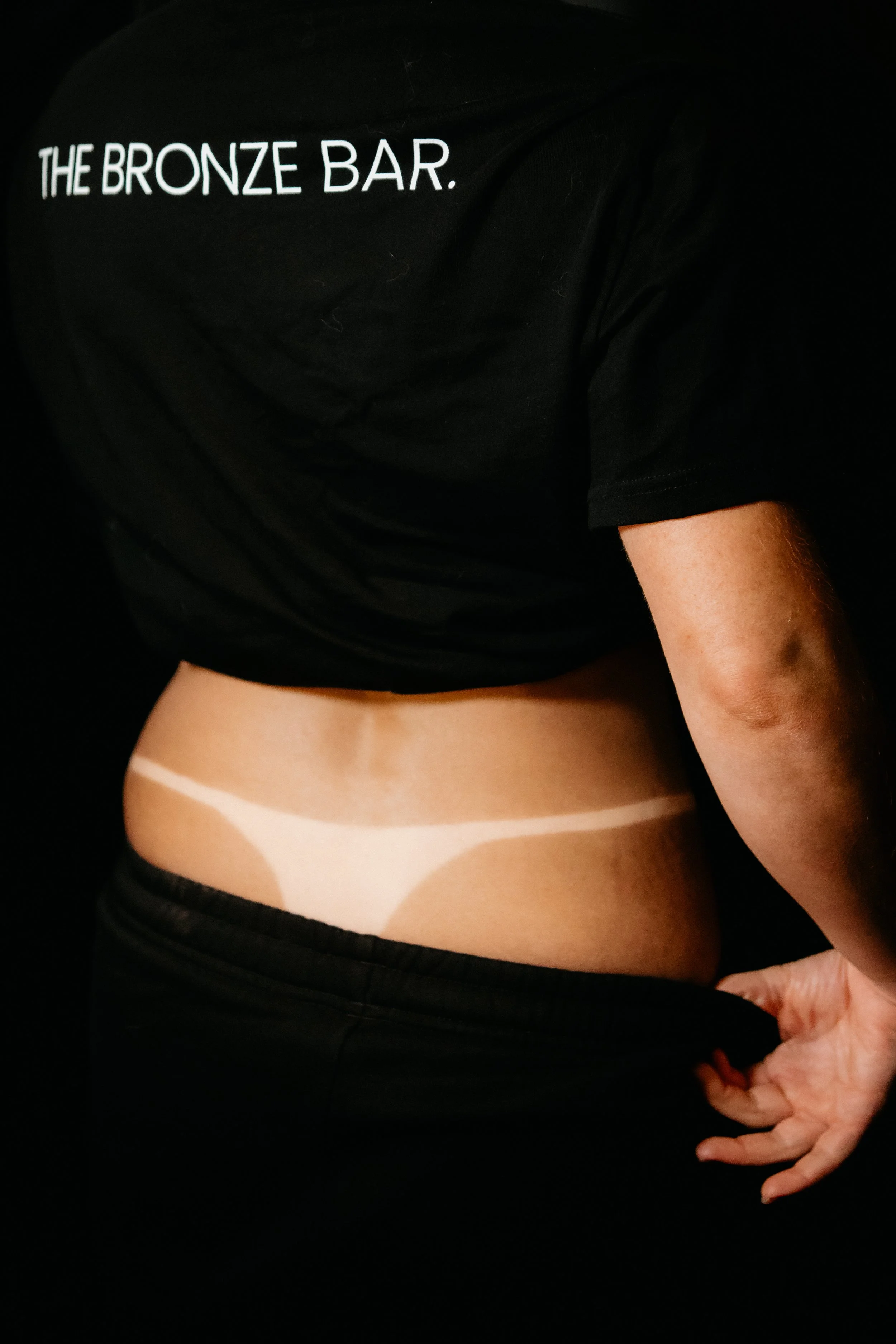 Person wearing a black T-shirt with 'The Bronze Bar.' written on it, lifting the shirt to reveal white body paint or markings on the lower back and waist area.