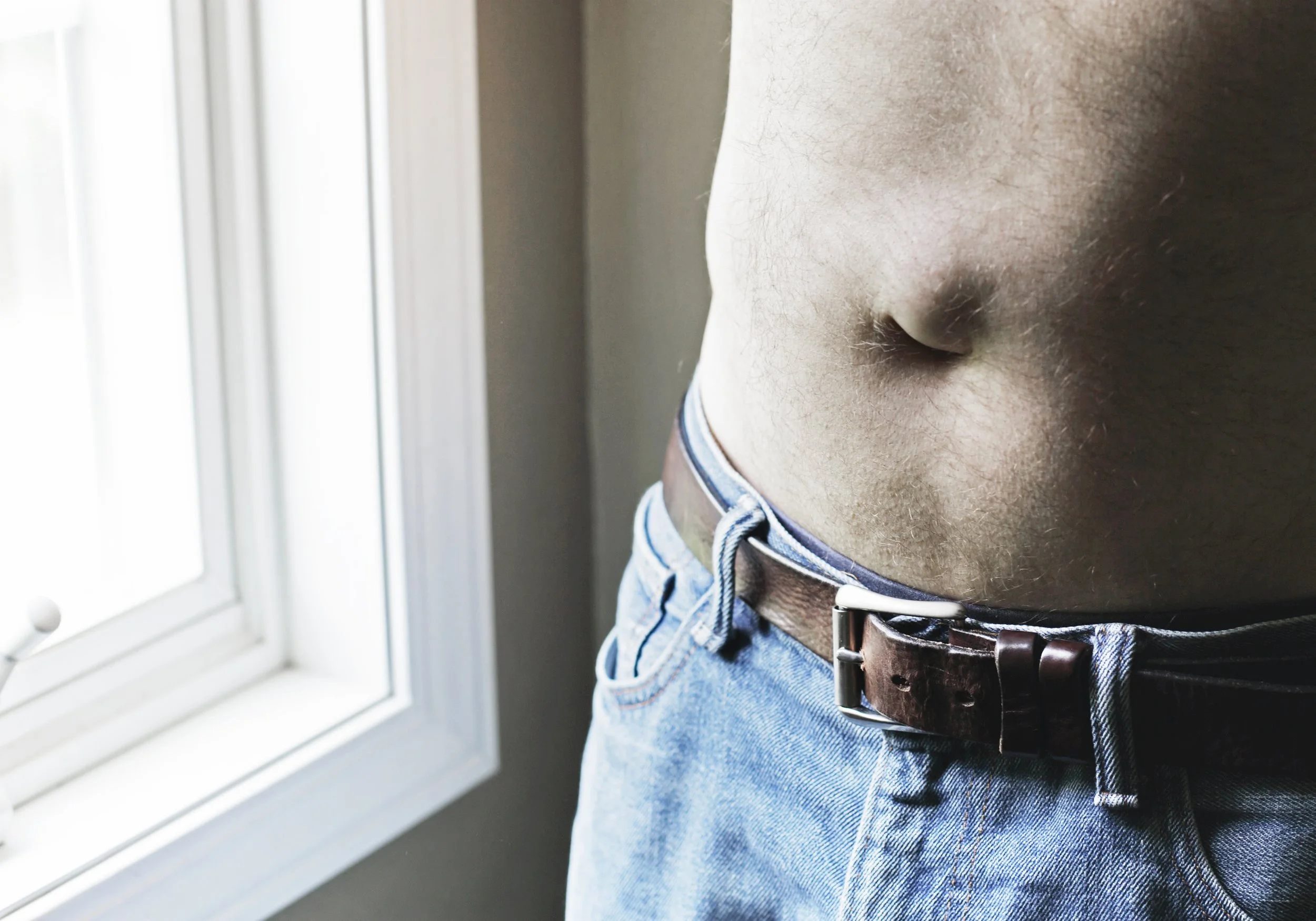 Close-up of a man's bare torso with umbilical hernia wearing blue jeans with a brown leather belt, standing near a window.