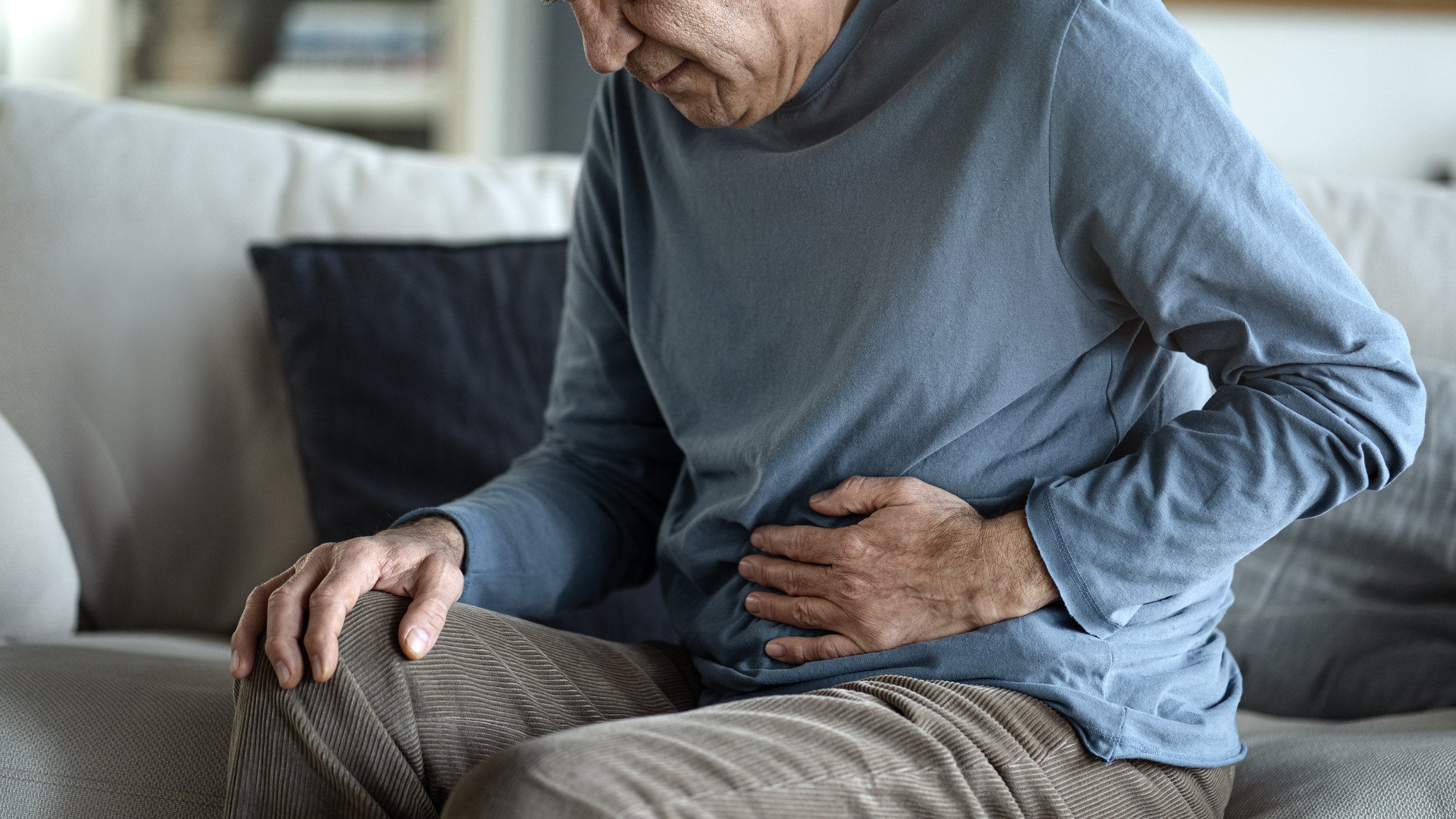 An elderly man sitting on a couch, holding his stomach with one hand and resting his other hand on his knee, indicating stomach pain or discomfort.