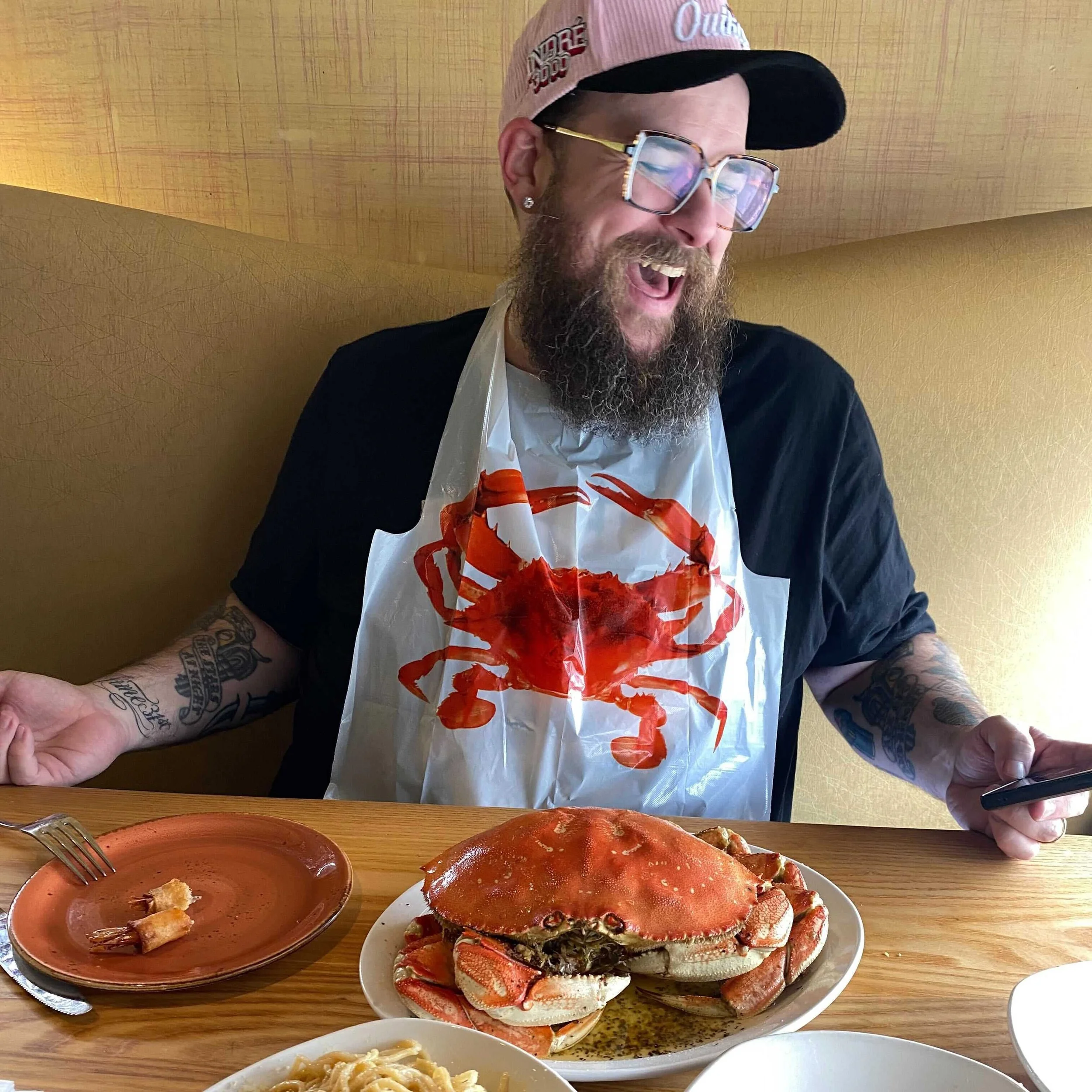 A man with glasses, a beard, and tattoos, wearing a pink baseball cap and a bib with a red crab on it, is sitting at a table enjoying a meal of crab and pasta.