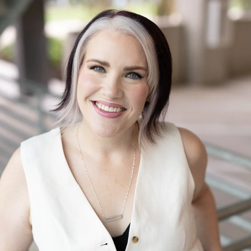A smiling woman with short, two-toned hair, wearing a sleeveless white top and a silver necklace, indoors with blurred background.