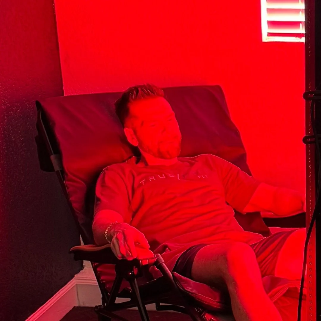 A man with short hair and a beard is reclining on a black chair possibly in a medical or relaxing setting, surrounded by red lighting.