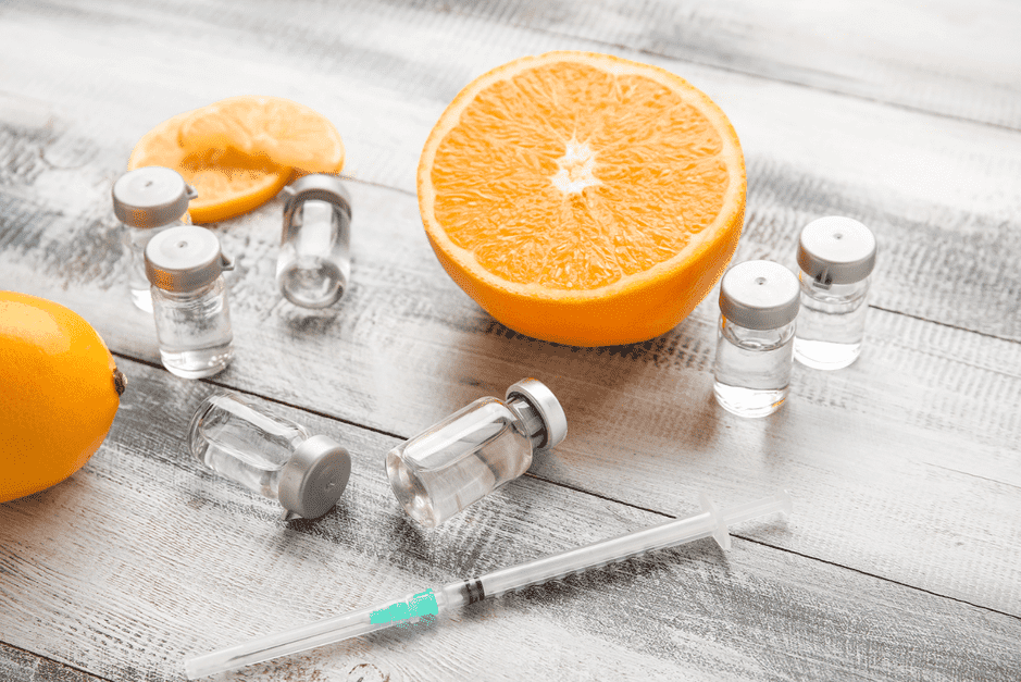Glass medicine vials and syringe next to halved orange and lemon on white wood background, illustrating vitamin C