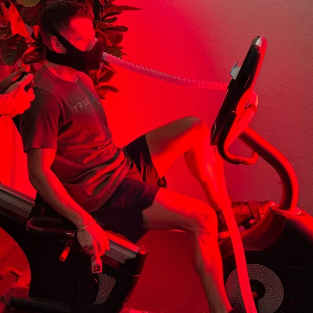 A woman in exercise gear undergoing a red light therapy session on a treadmill, which she is sitting on, with a tube connected to her face.