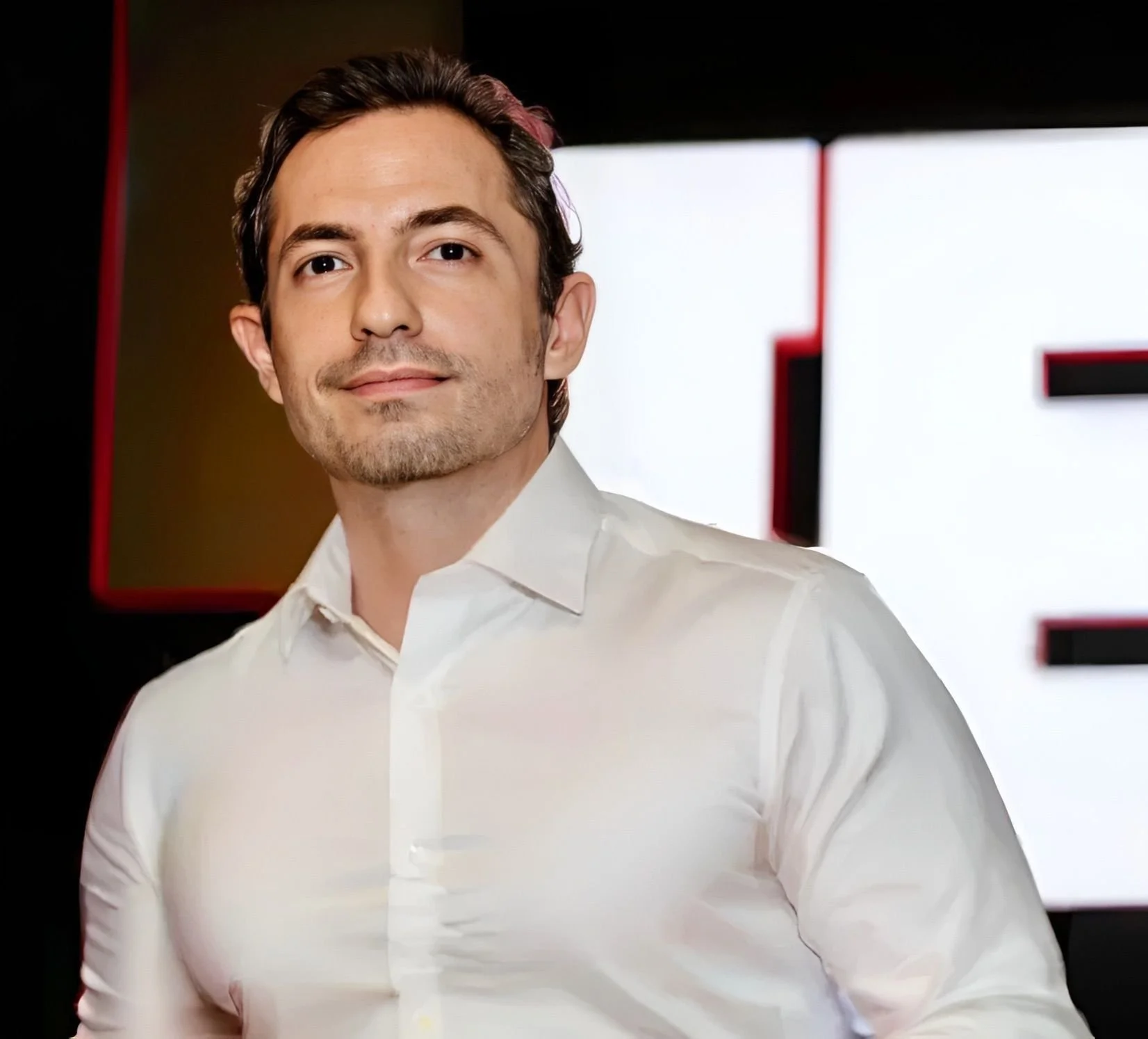 A young man with dark hair and a light beard, wearing a white dress shirt, standing in front of a background with large white and black shapes.